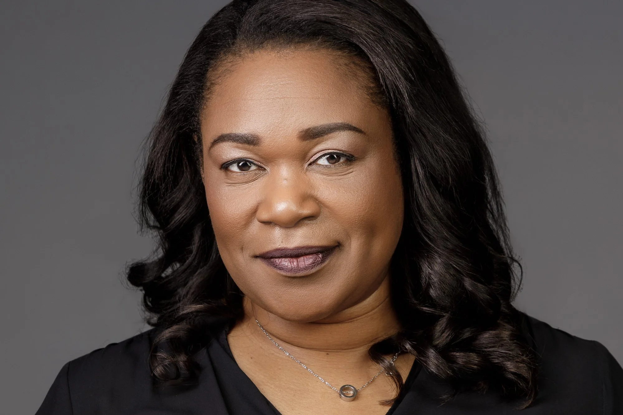 Portrait of a business woman/person with long black hair, wearing a black top and a necklace, against a gray background.