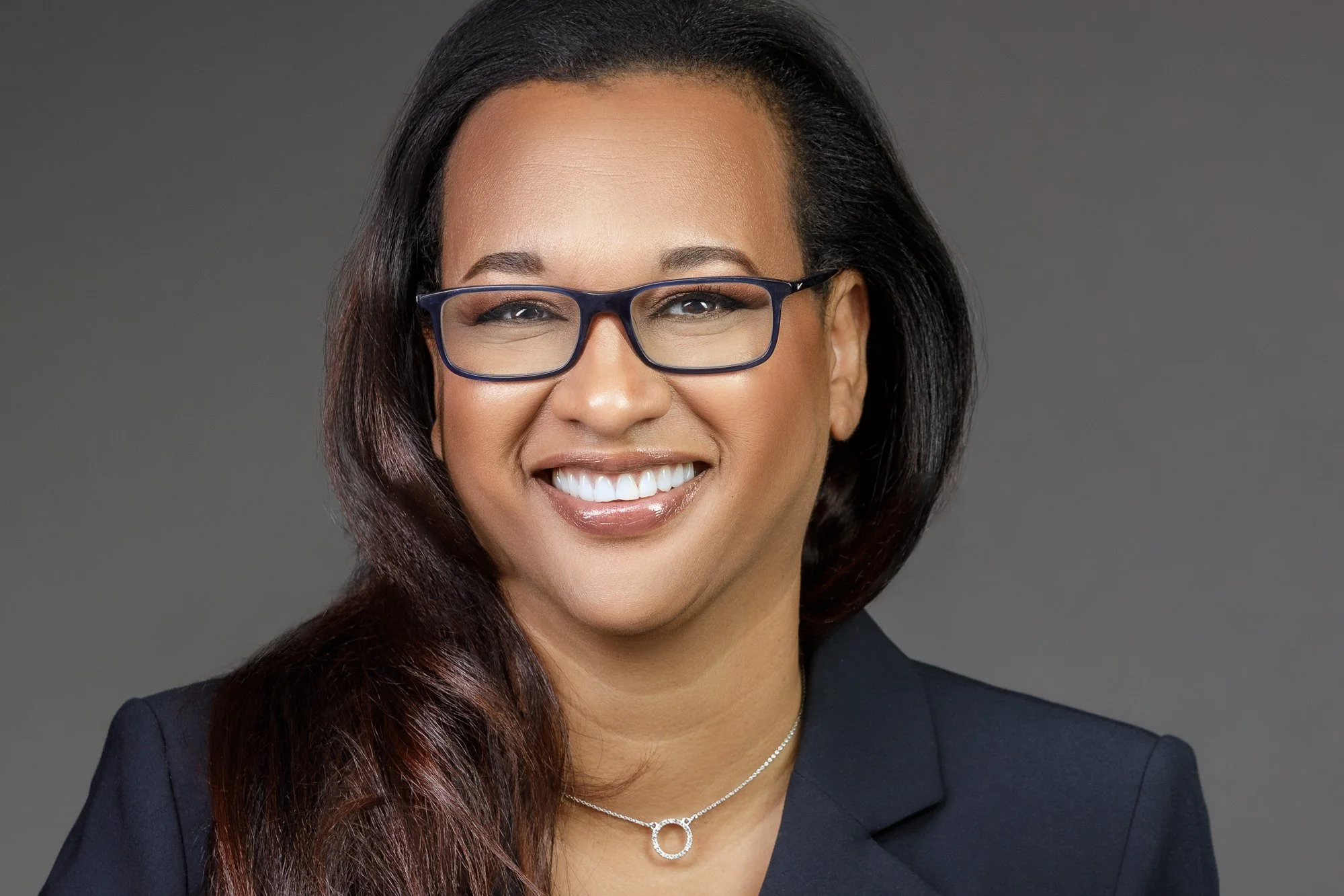 Portrait of a professional business woman smiling wearing glasses and a necklace, dressed in black suit against a gray background.