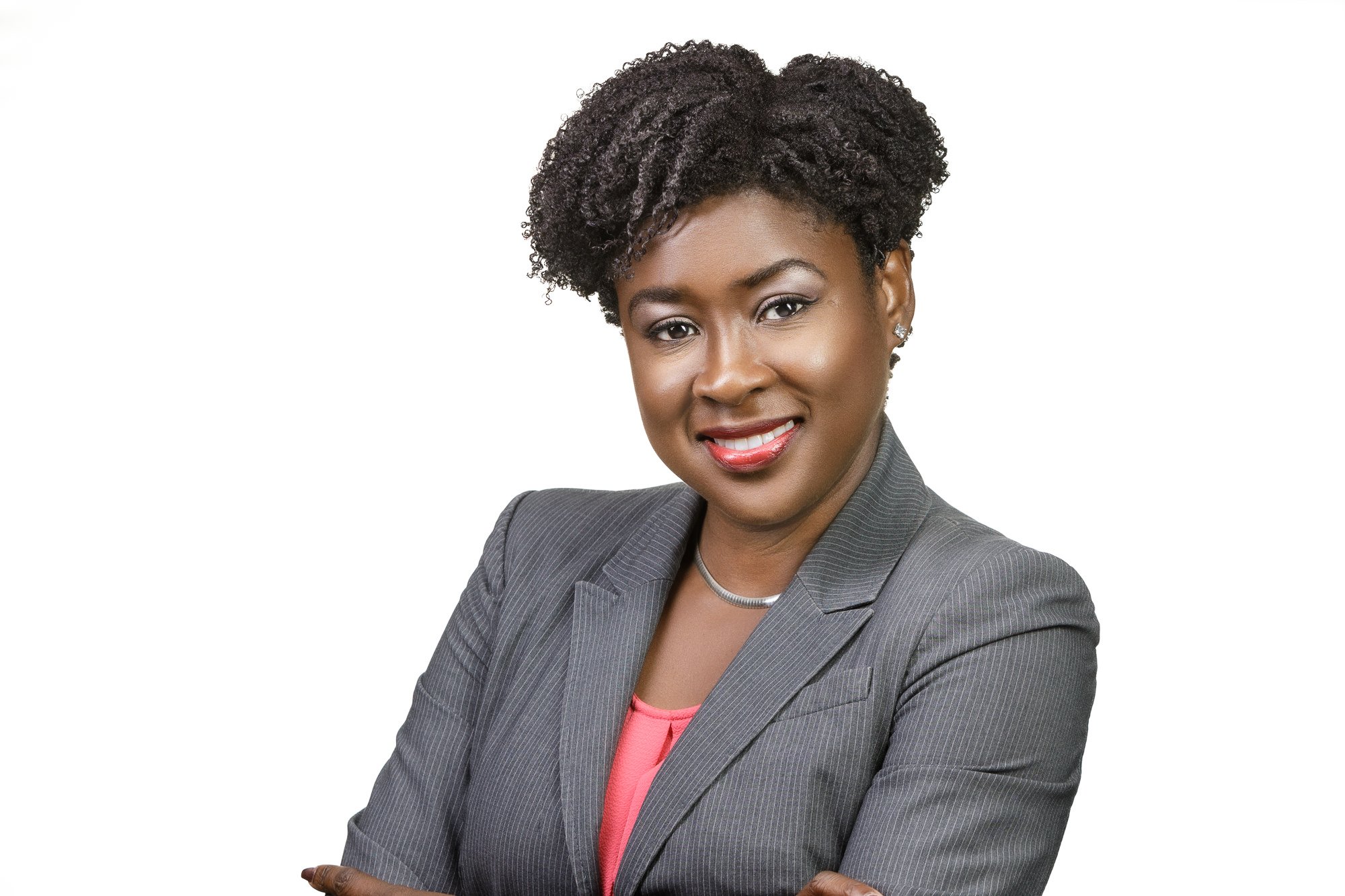 Professional woman in a gray suit with natural hair, smiling and crossing arms against a white background.