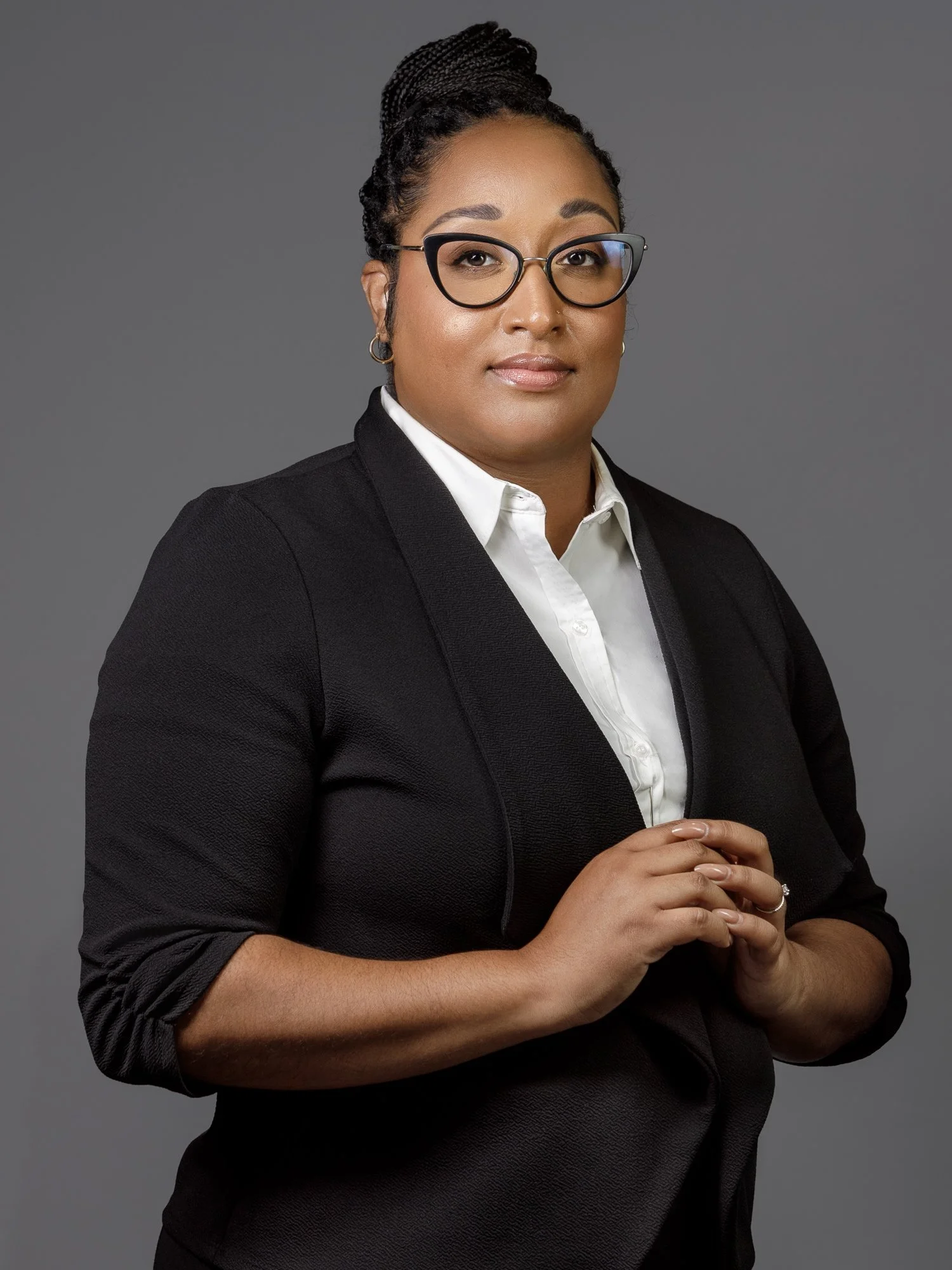 A business person wearing glasses and a black blazer over a white shirt, standing against a gray background, with hands clasped.