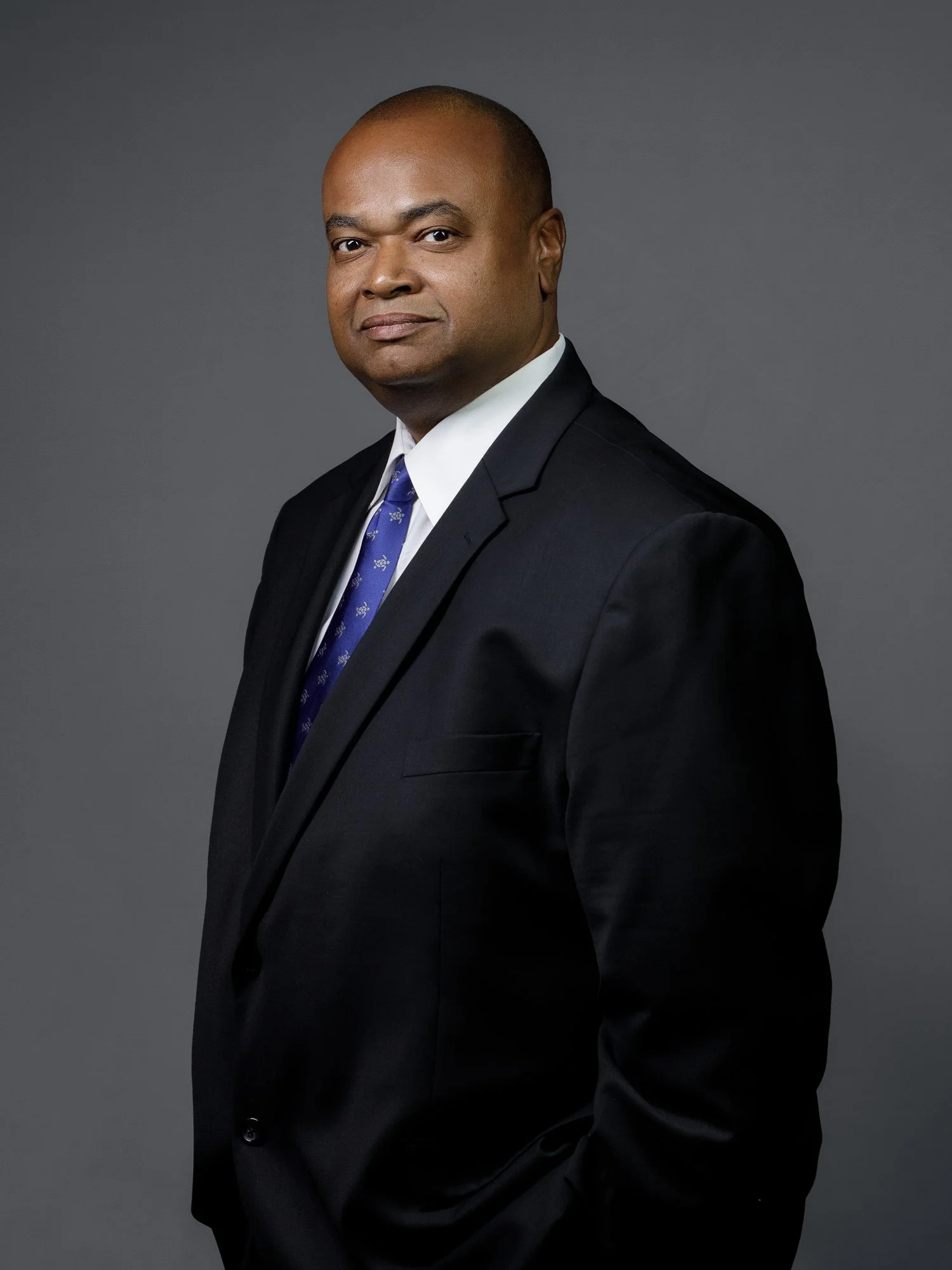 Man in a black suit with a blue tie posing against a gray background.