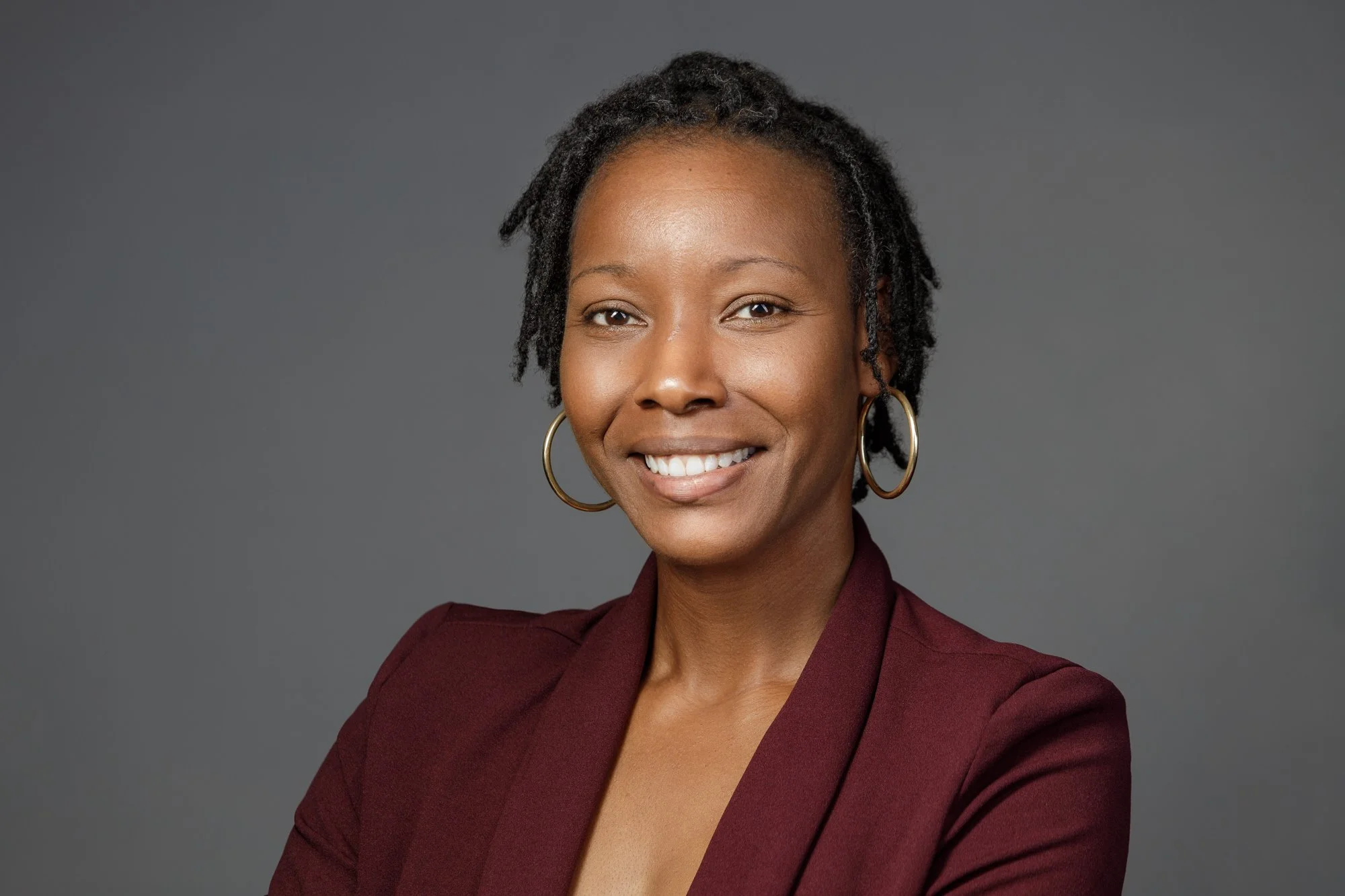 Smiling woman with short natural hair, wearing a burgundy blazer and gold hoop earrings against a gray background.