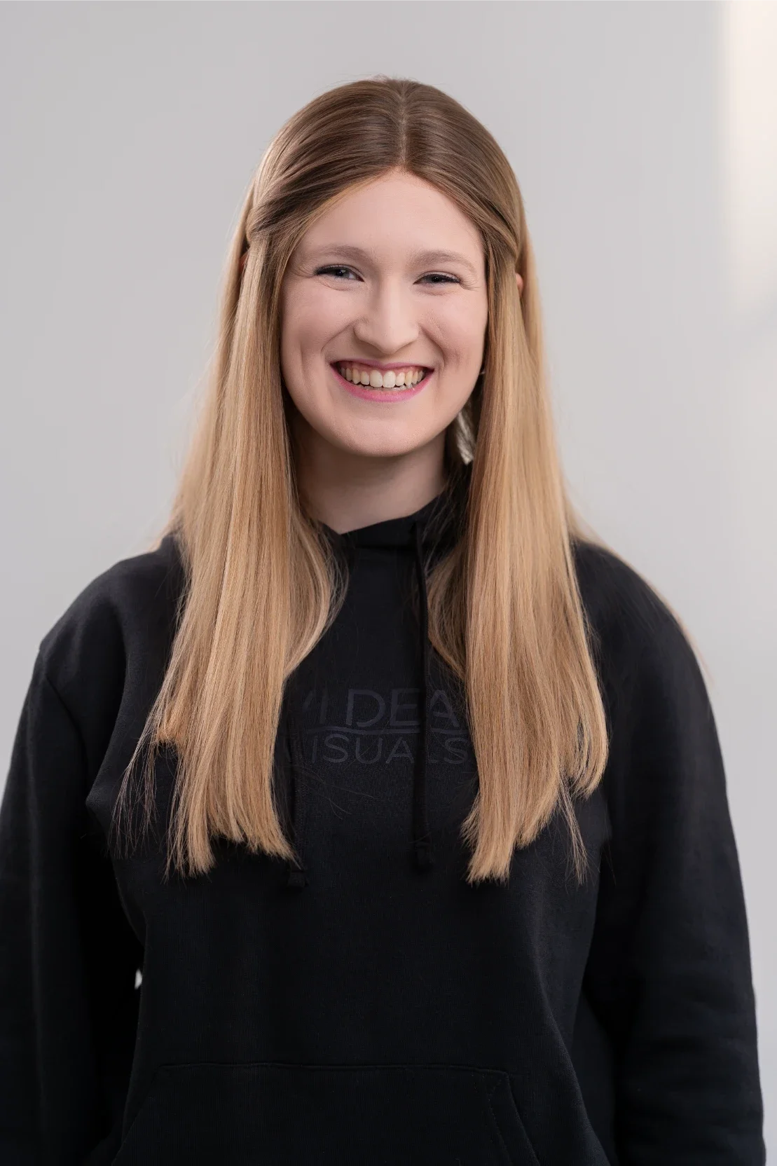 A smiling woman with long, straight ginger hair wearing a black hoodie standing against a plain light gray background.