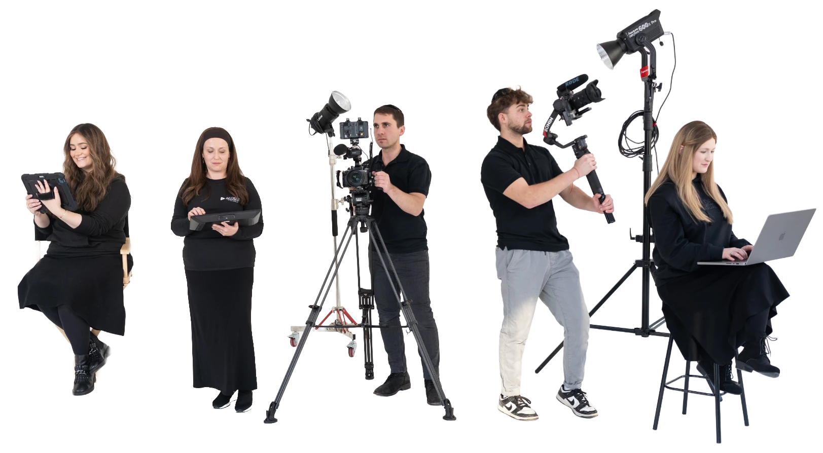 A filming crew working on a production set with five people: a woman sitting on a chair reading a tablet, a woman standing and holding a slate, a man operating a camera on a tripod, a man holding a large light on a stand, and a woman sitting on a stool using a laptop.