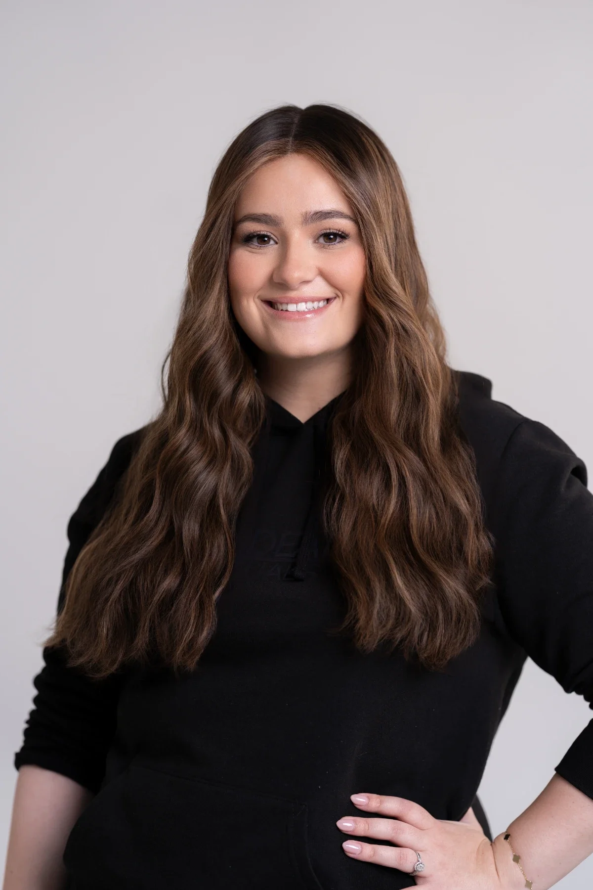 A woman with long, wavy brown hair, smiling, wearing a black hoodie, standing against a plain light gray background.