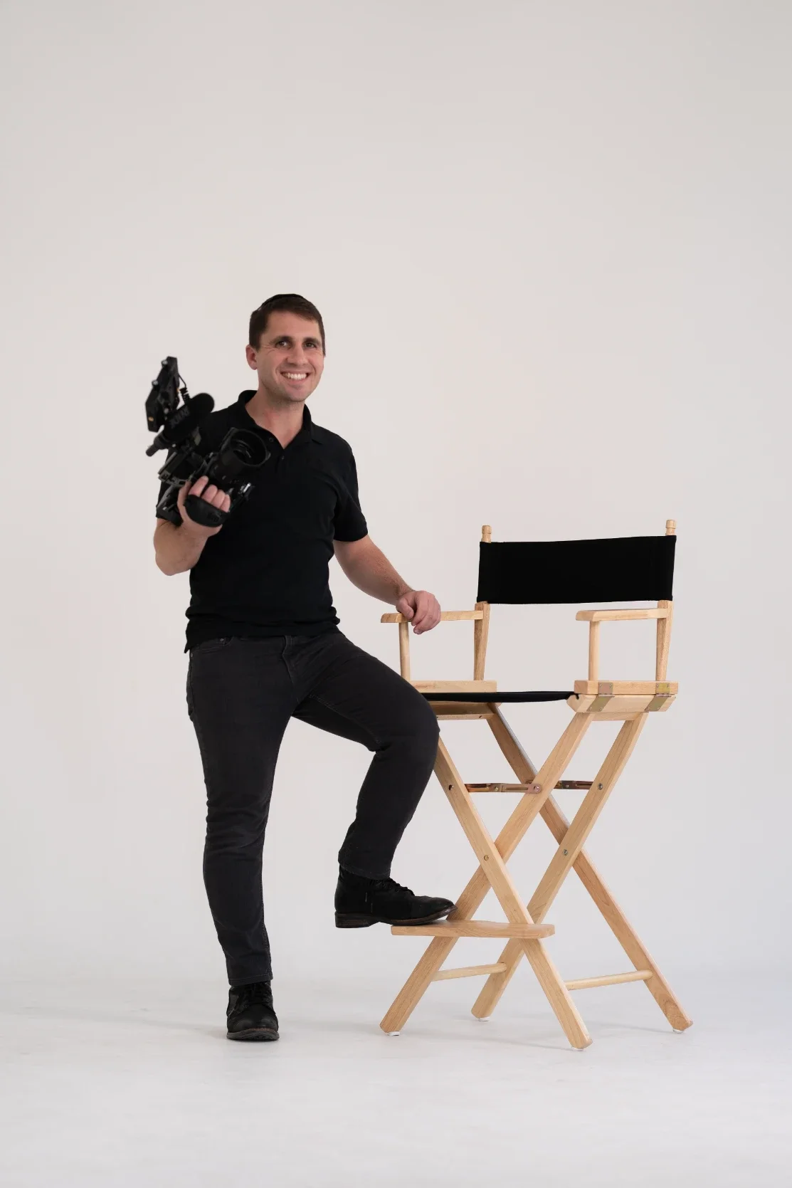 A man dressed in black holding a camera, standing next to a director's chair with a black seat and backrest against a plain white background.