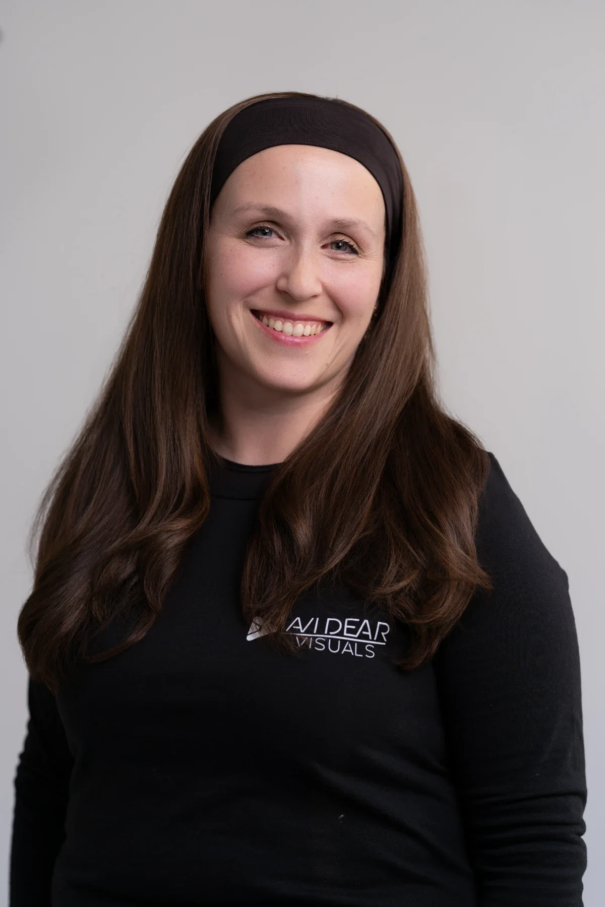 A woman with long brown hair wearing a black headband and black shirt smiling against a plain gray background.