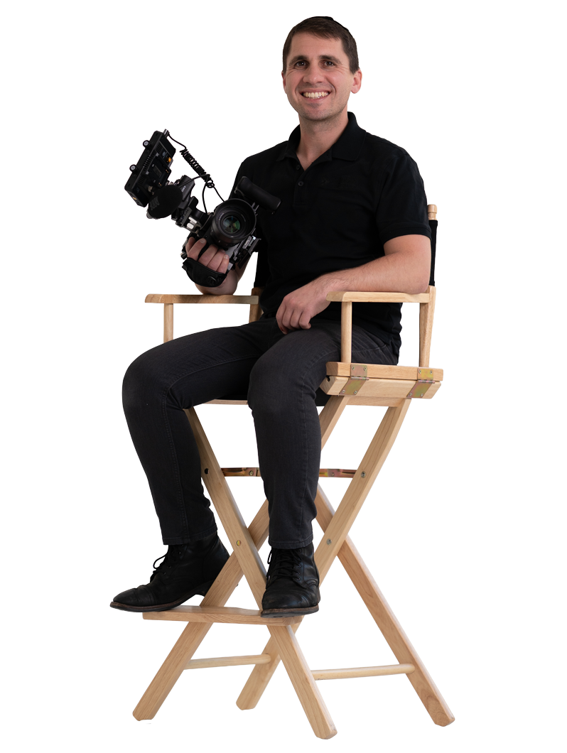 A man sitting on a wooden director's chair holding a professional video camera, smiling at the camera.