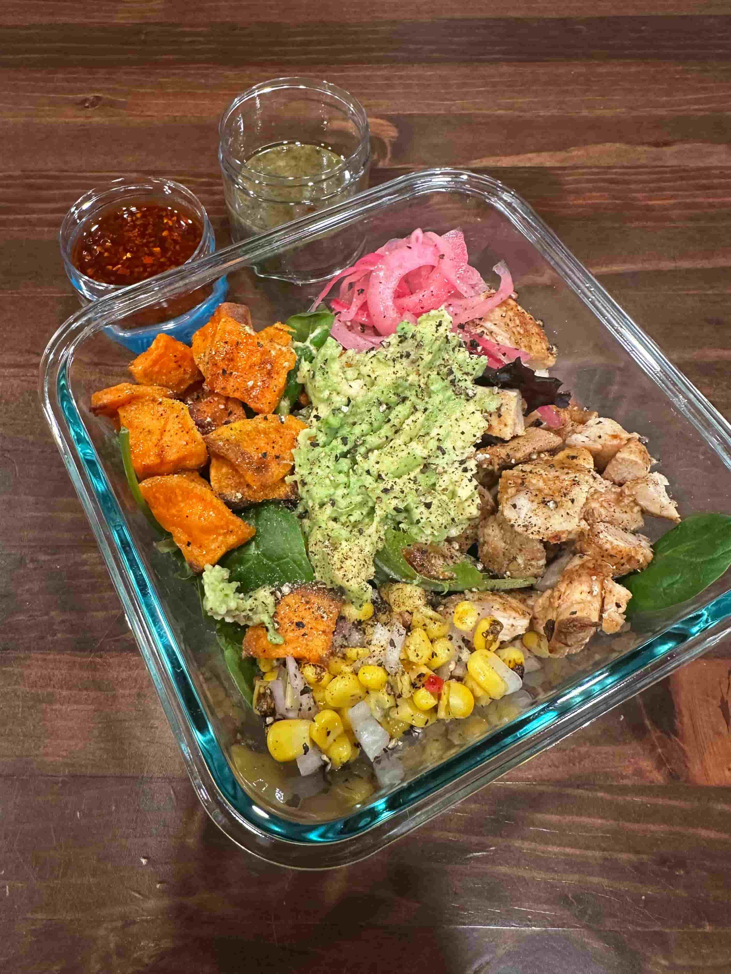 Healthy batch prep chicken salad bowl in glass meal prep container with grilled chicken, mashed avocado, roasted sweet potato, corn salsa, pickled onions and dressing on wooden table.