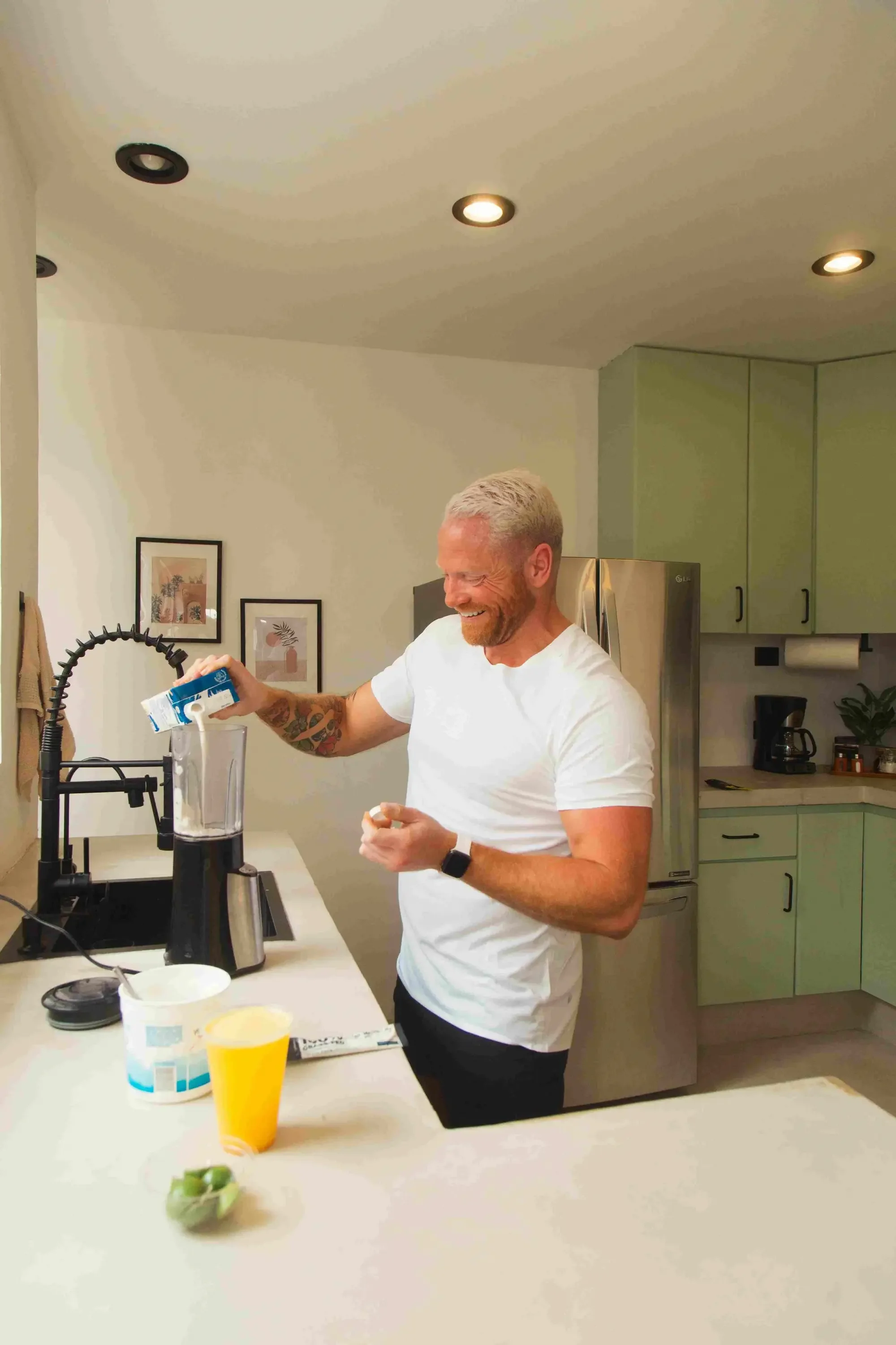 Coach Zach, in a kitchen, pouring milk from a carton into a blender. He is wearing a white t-shirt and a smartwatch. The kitchen has green cabinets, a stainless steel refrigerator, and framed artwork on the wall.