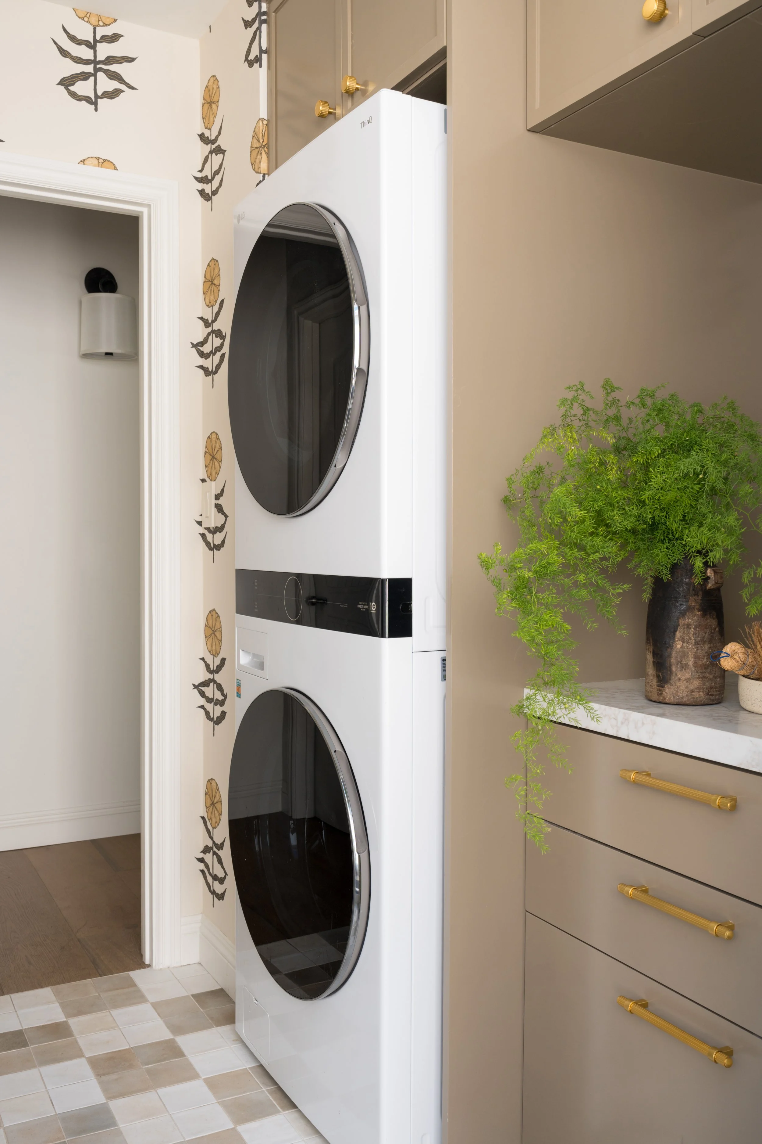 Stacked white front-loading washer and dryer in a laundry room with beige cabinetry and a potted plant on a white countertop.