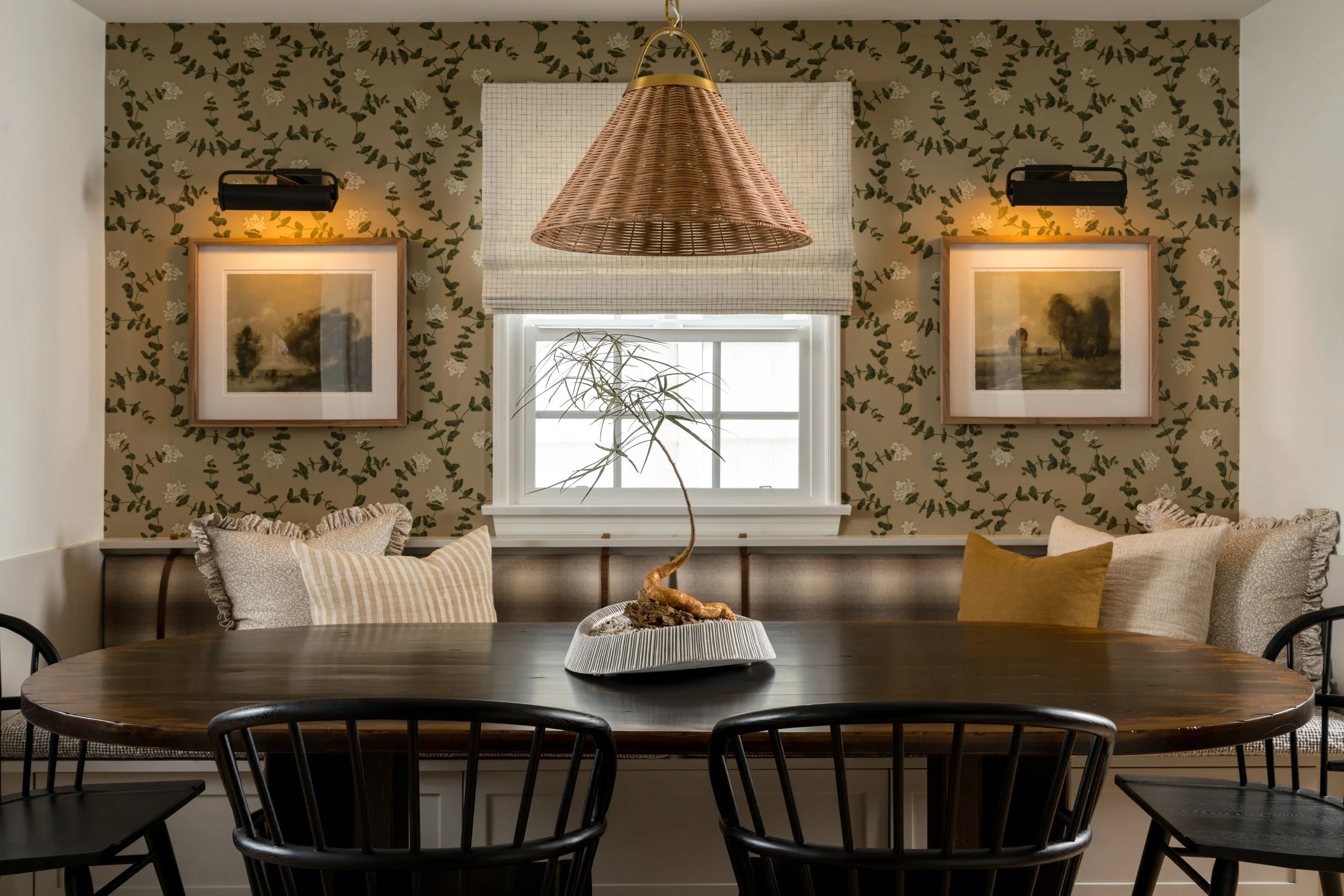 A cozy dining nook featuring a dark wooden oval table with black wooden chairs. The background has a window with a white frame, floral wallpaper, and two framed landscape paintings with warm lighting. There are beige, striped, and brown throw pillows on the built-in bench seating. A paper plant in a decorative container sits on the table, and a rattan pendant light hangs above.