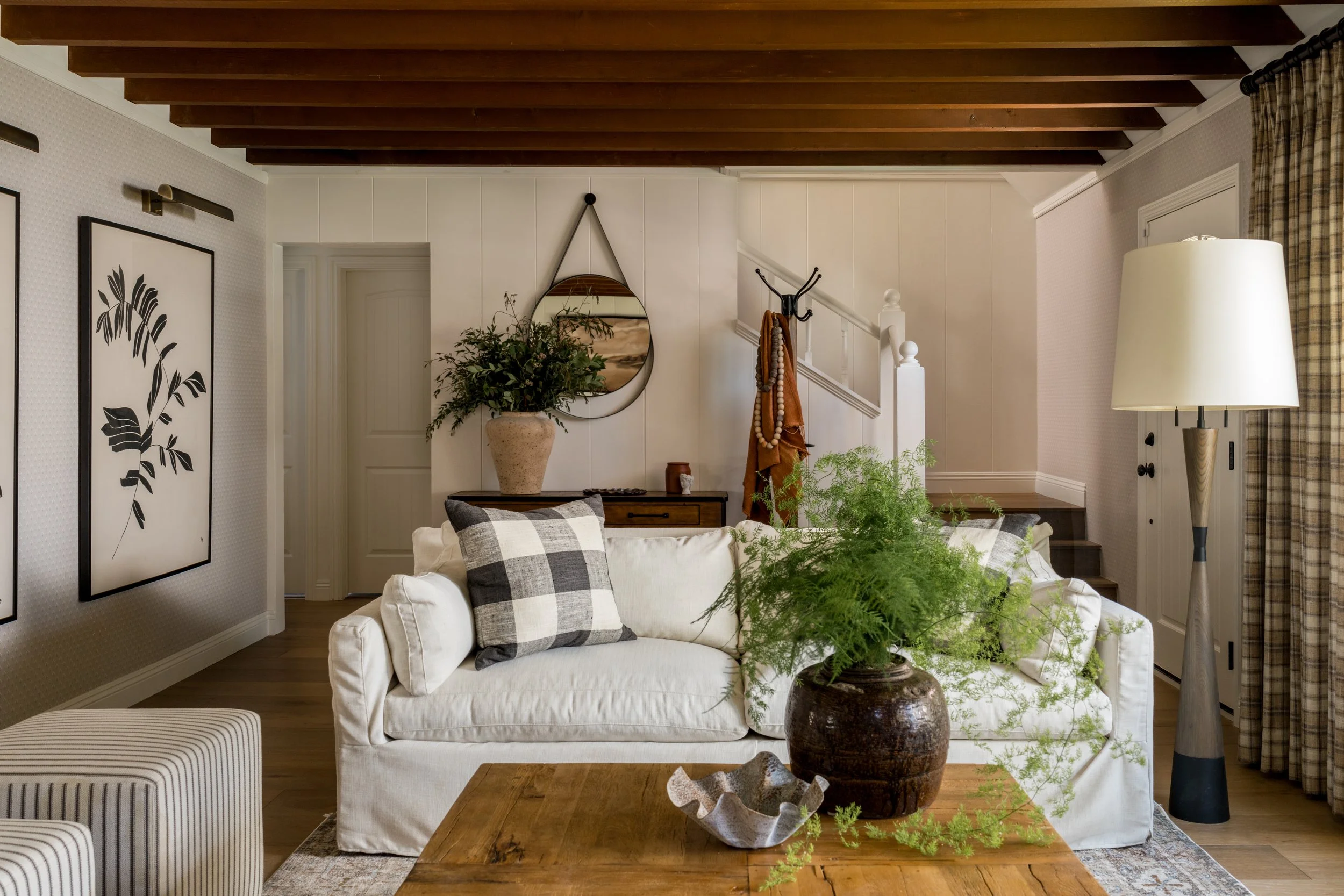 Living room with white sofa, checkered pillows, wooden coffee table with a large potted fern, floor lamp, wall art, and a staircase with a coat hook and plants in a homey, well-decorated space.