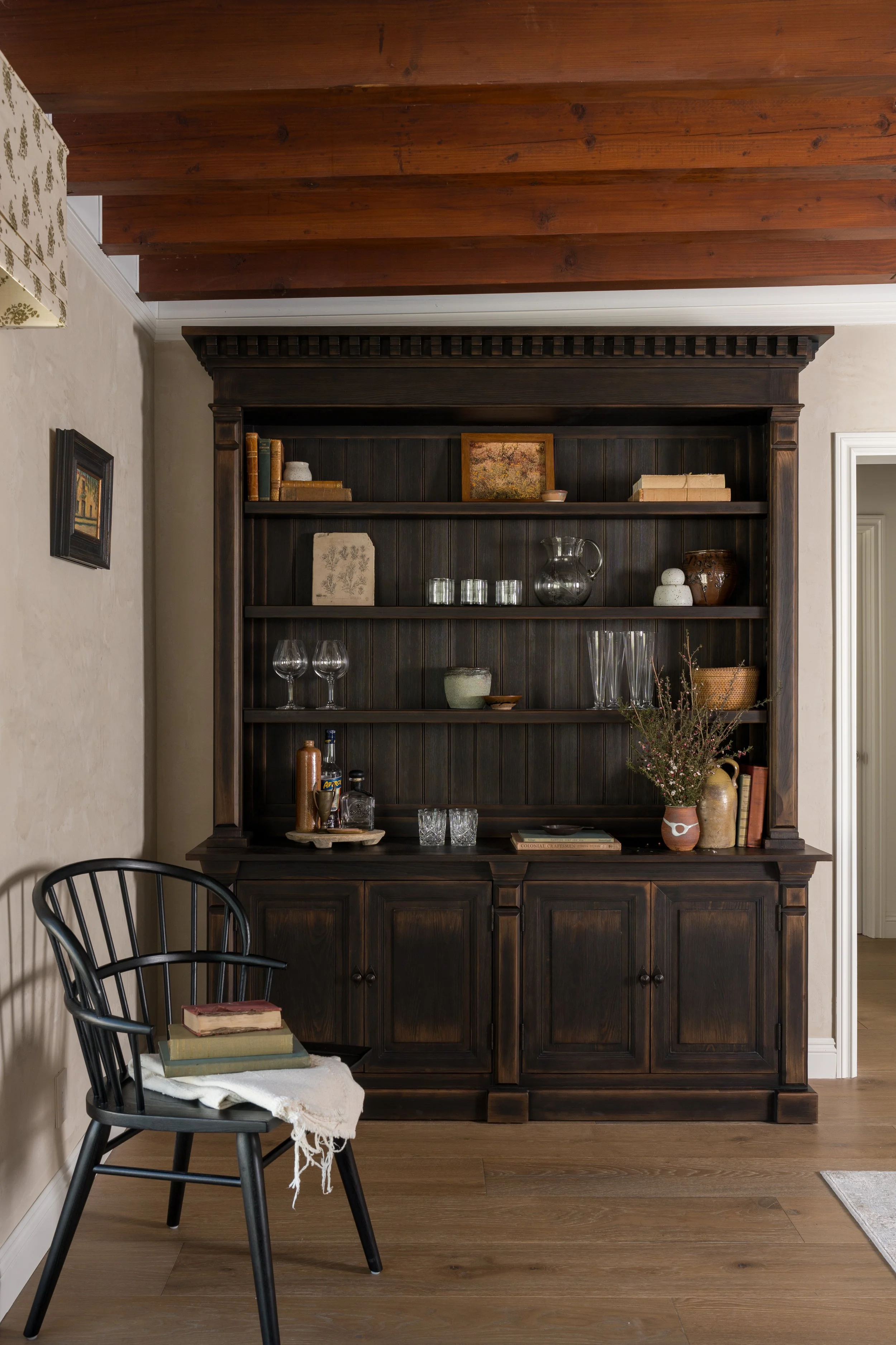 A dark wooden china cabinet filled with glassware, books, and decorative items, against beige walls and a wooden ceiling, with a black chair and stacked books in the foreground.