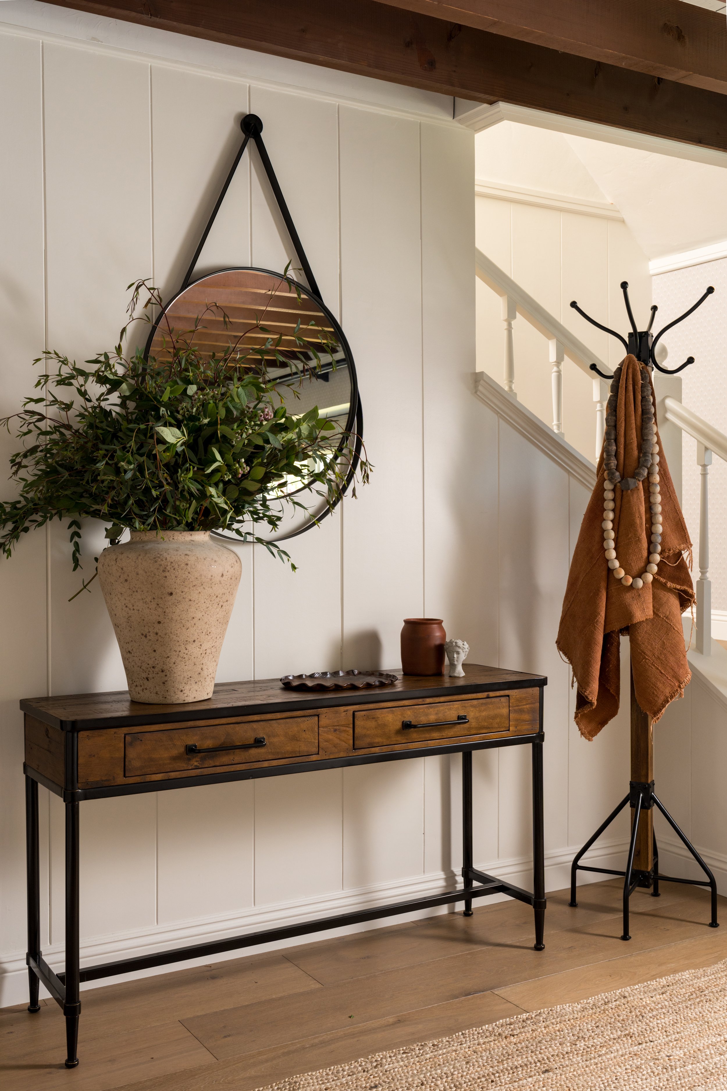 A hallway corner with a black metal and wood console table, large vase with greenery, oval mirror hanging from a black strap, and a coat stand with brown towels and beaded necklaces.