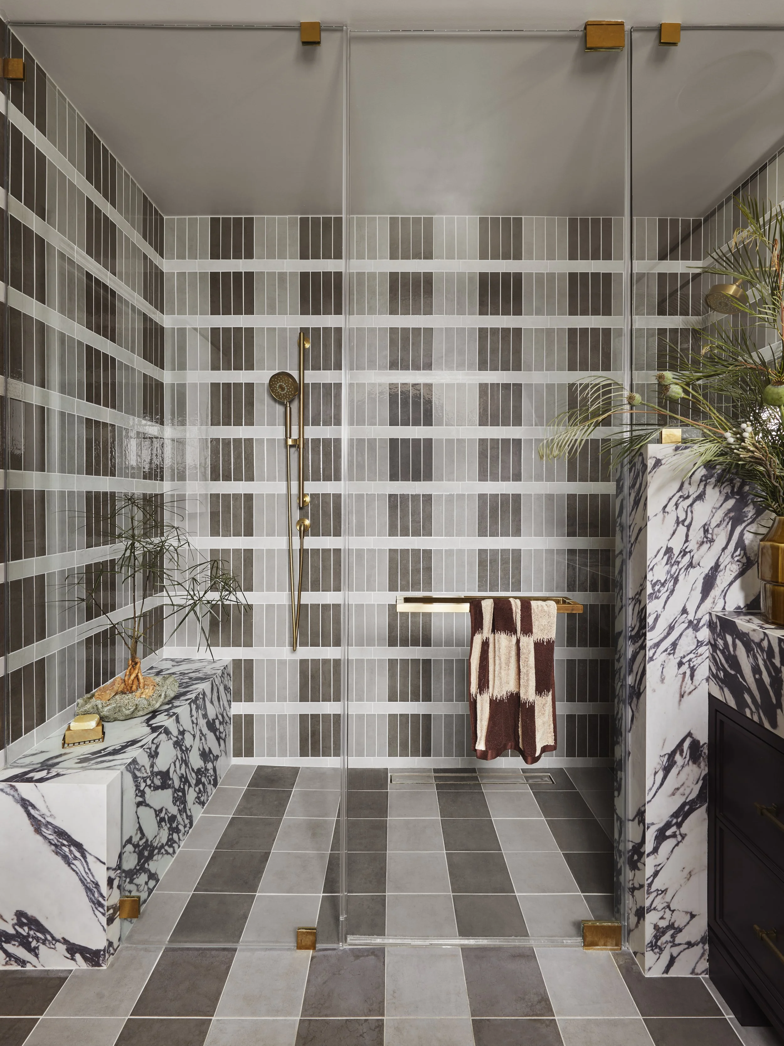 A modern bathroom with a glass shower enclosure, gray and white tiled walls, and marble accents. Decorative plants and a towel hang on a brass towel bar.