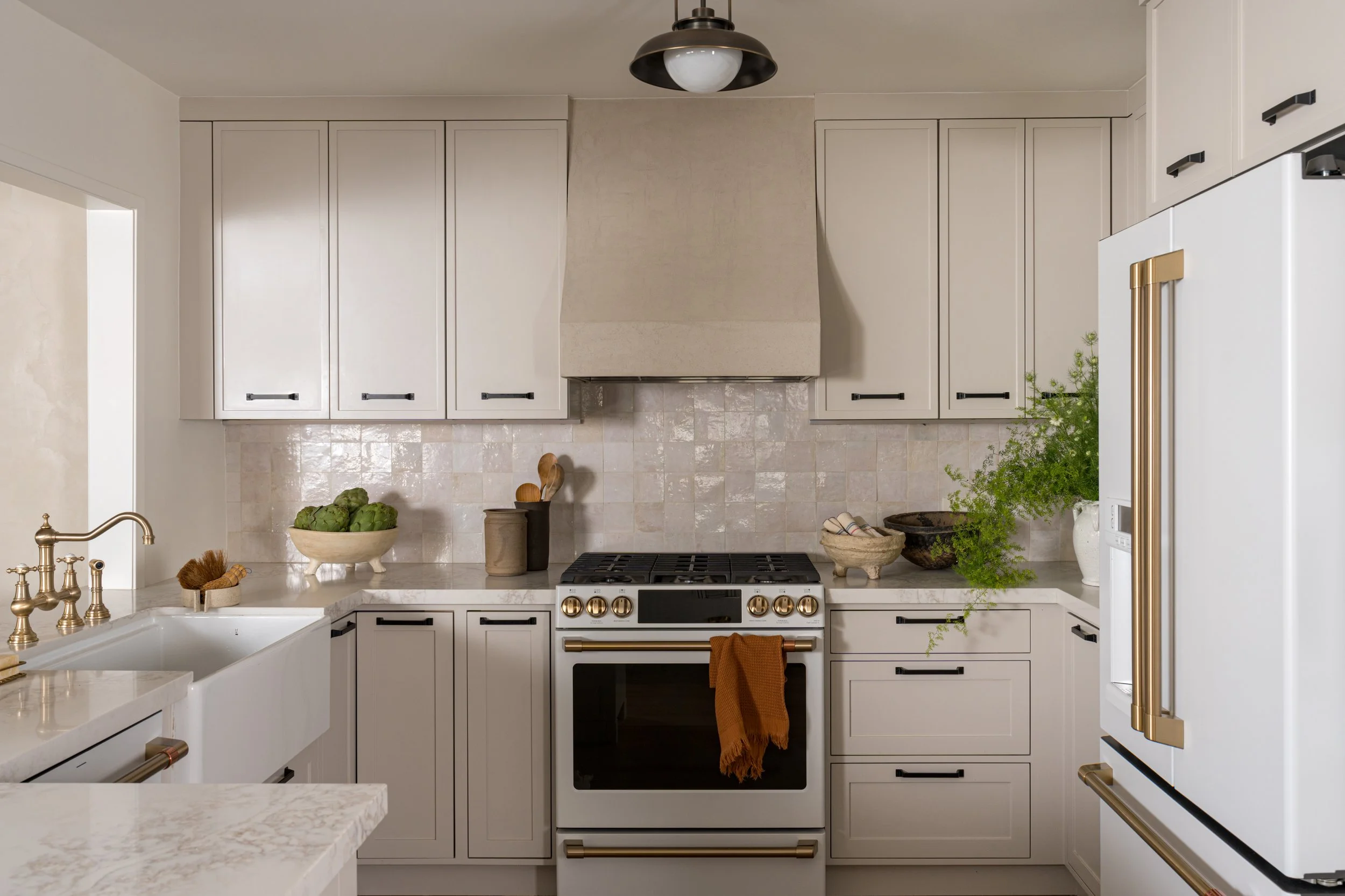Modern kitchen with white cabinets, gold hardware, stainless steel stove, and a cream-colored backsplash. Decor includes a bowl of artichokes, plants, and kitchen utensils.