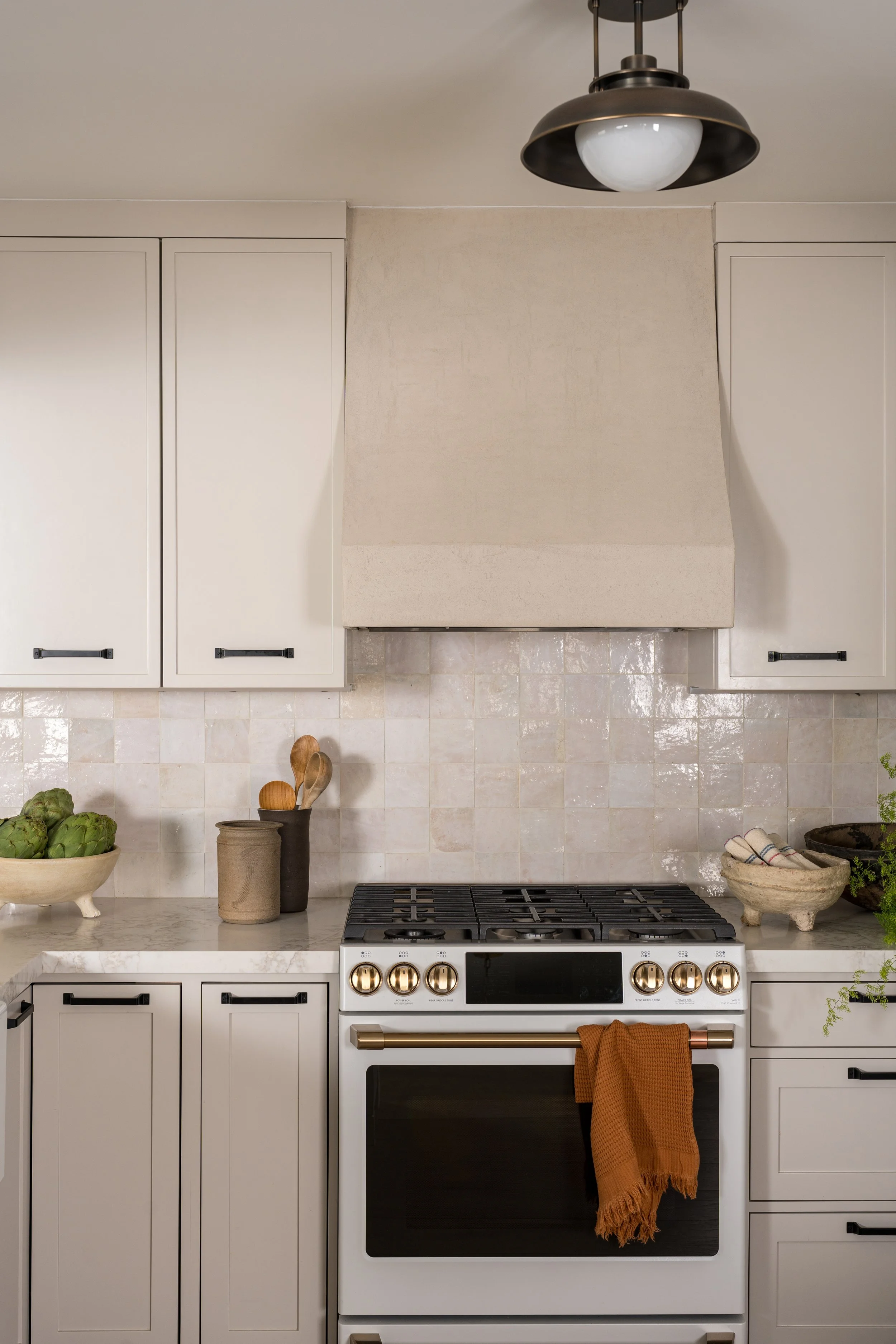 Kitchen with white cabinets, a beige range hood, a gas stove with gold knobs, and a white oven. Decor includes artichokes, wooden utensils, and a terracotta-colored towel hanging from the oven handle.