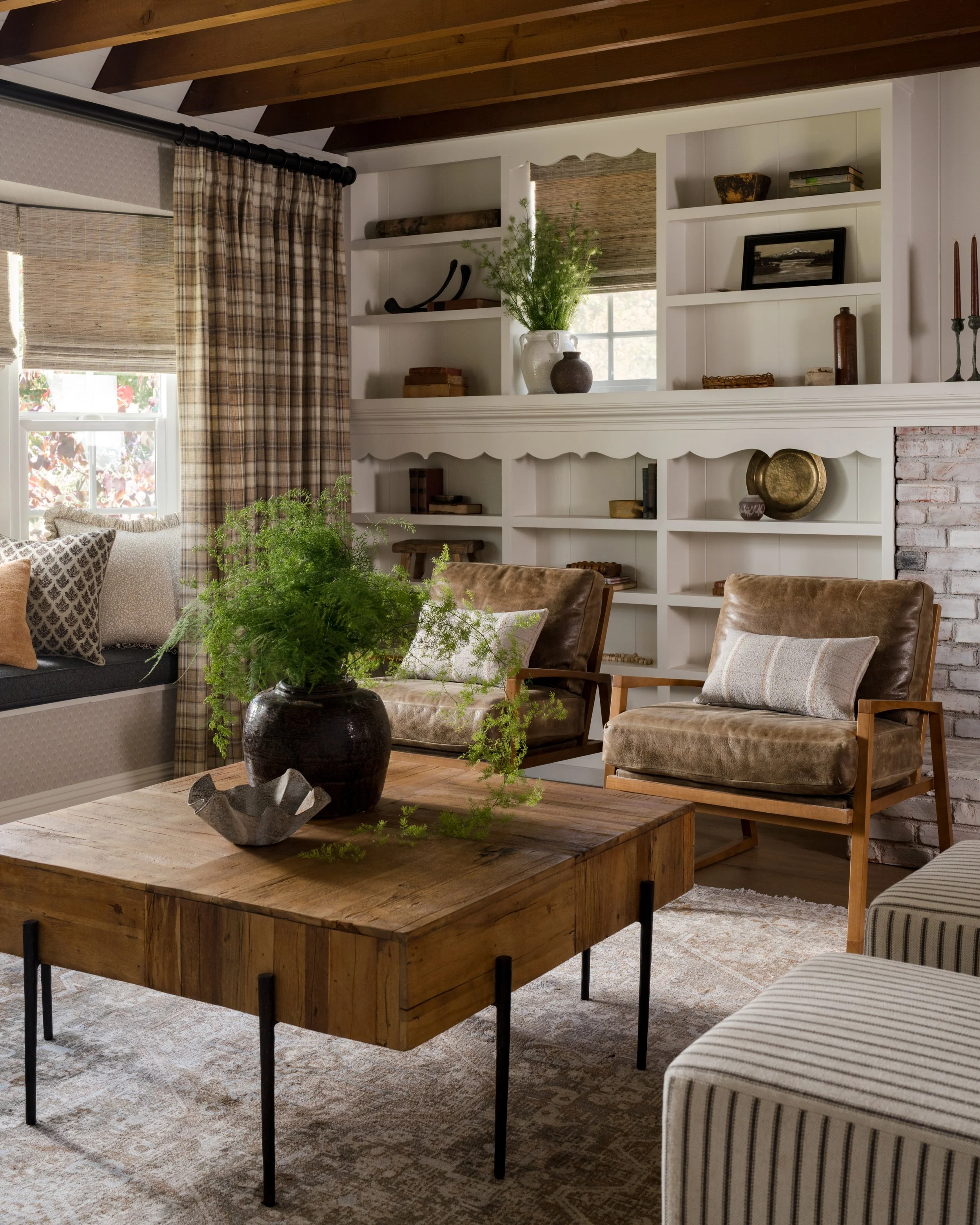 Living room with wooden ceiling, beige curtains, bookshelf, leather chairs, wooden coffee table, and decorative cushions.