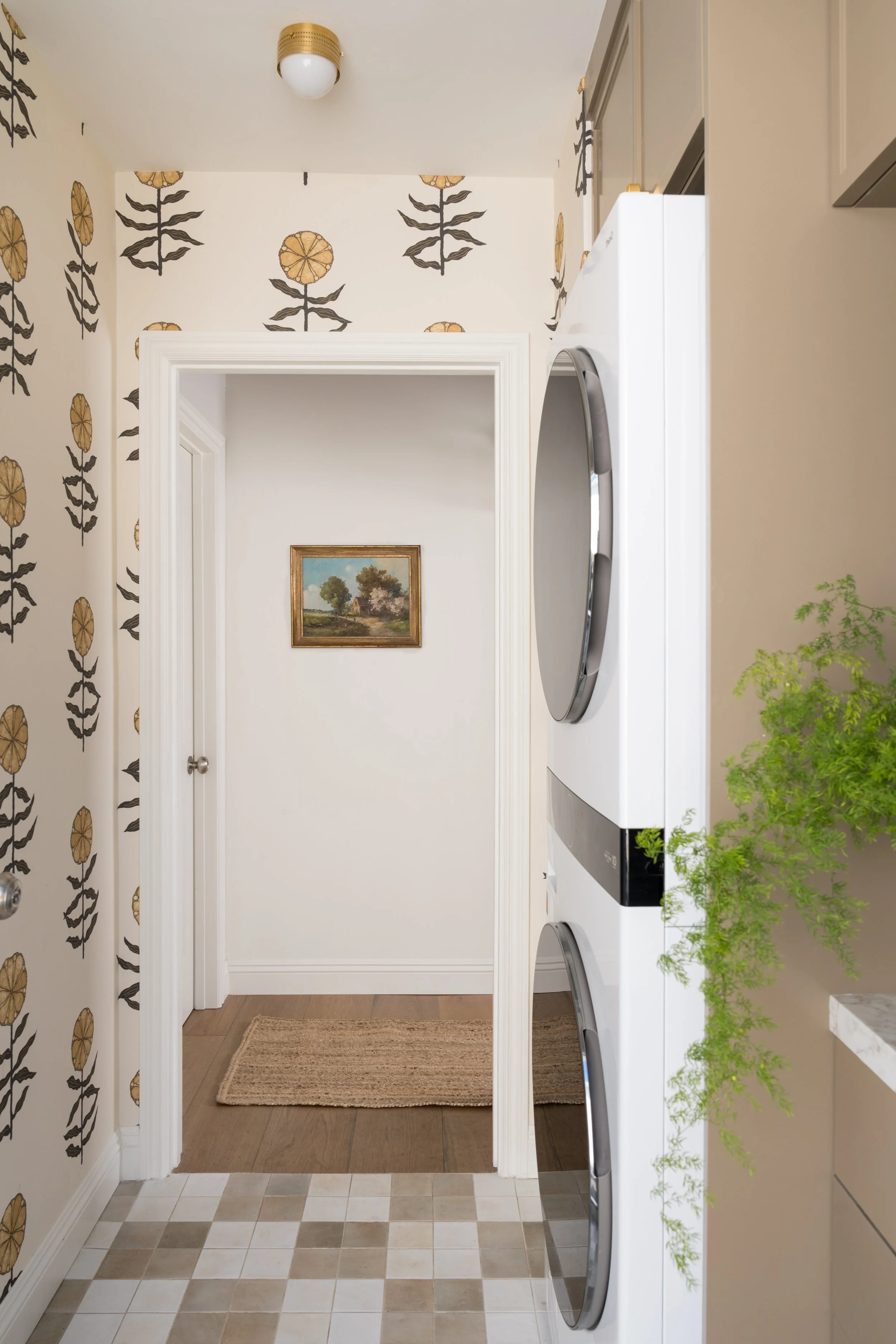 A laundry room with a stacked washer and dryer, beige cabinets, a green plant on the right, and a doorway leading to a hallway with wood flooring and a small framed landscape painting on the wall.