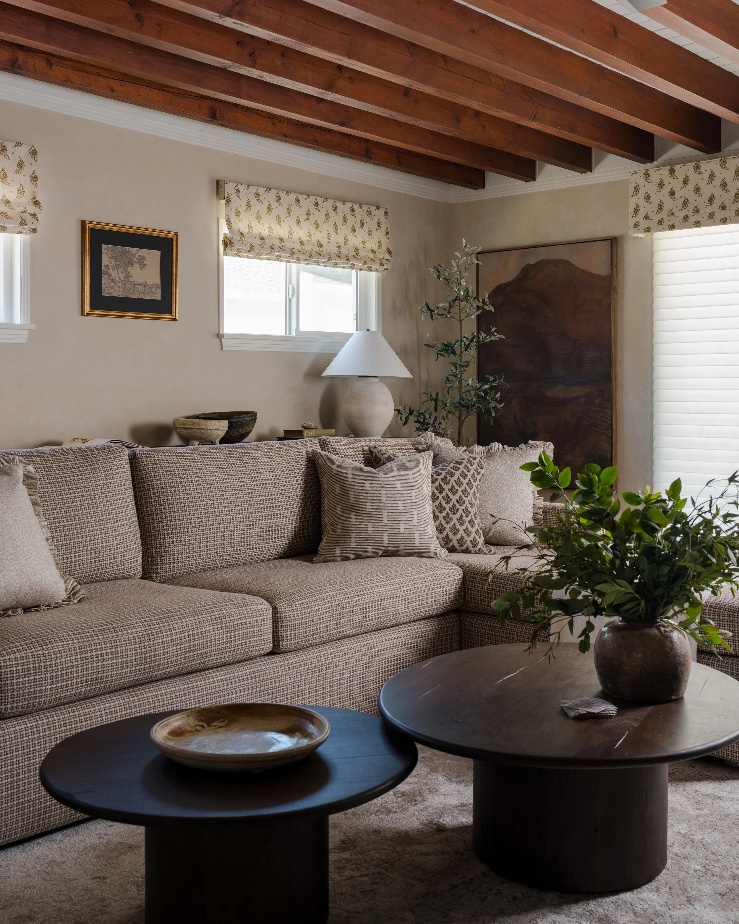 Living room with beige plaid sofa, decorative pillows, wooden ceiling beams, framed art, window blinds, potted plant, table lamp, and side tables with pottery and greenery.