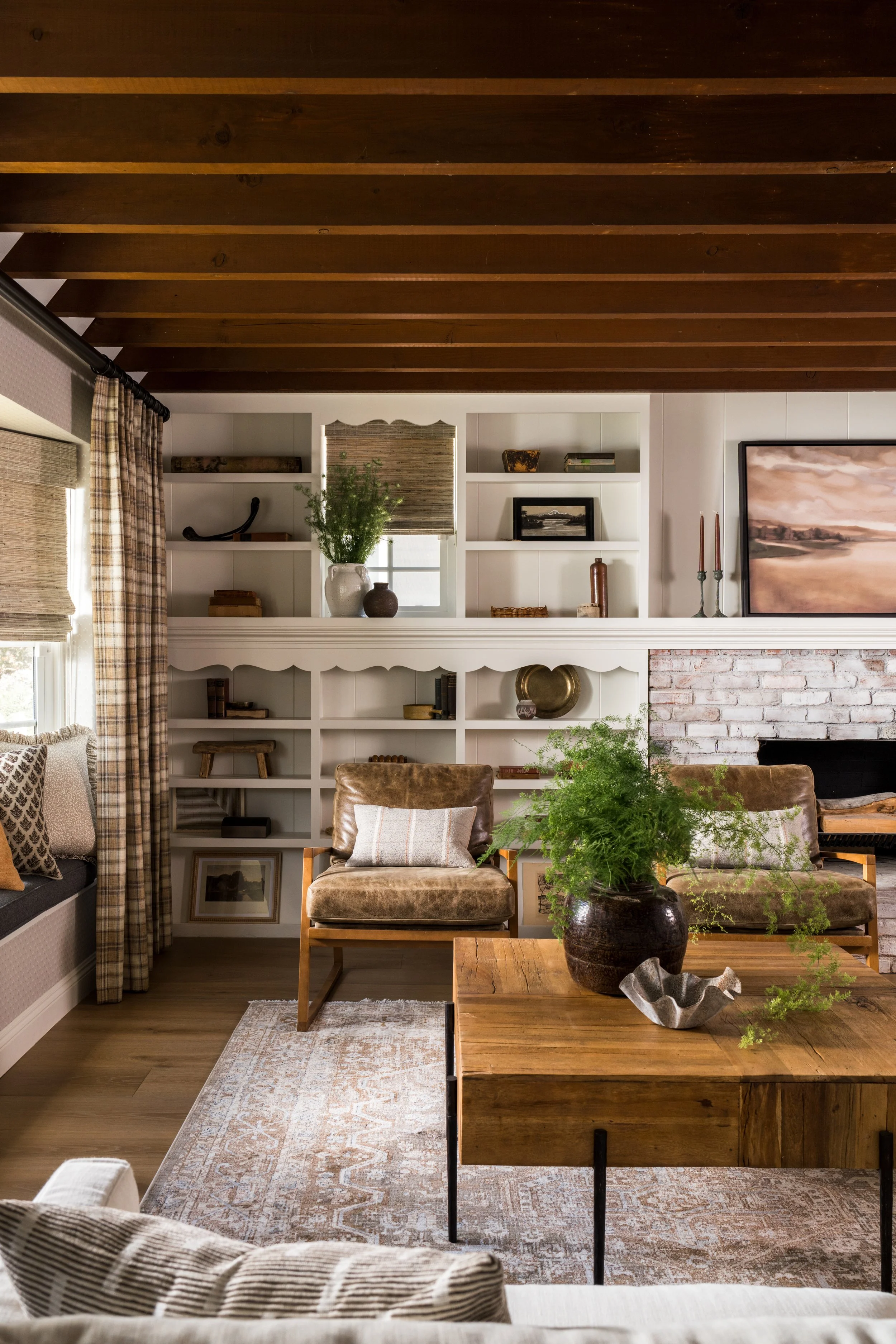 Living room with wooden ceiling, white built-in shelves, a brick fireplace, brown leather chairs with pillows, a wooden coffee table with a vase of green plants, patterned curtains, window seats, and artwork on the walls.