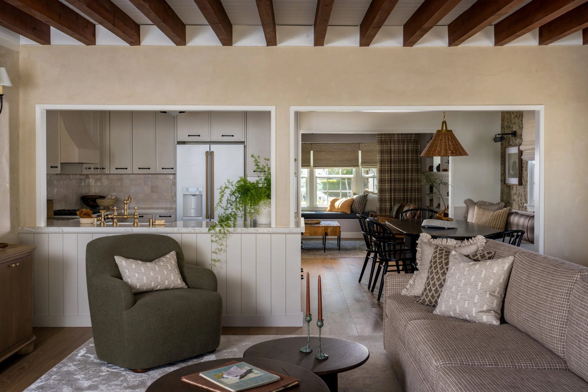 Living room with view into a kitchen and dining area, featuring a beige sofa, green armchair, wooden coffee tables, and a dining table with black chairs, bright window seat, and various decorative pillows.
