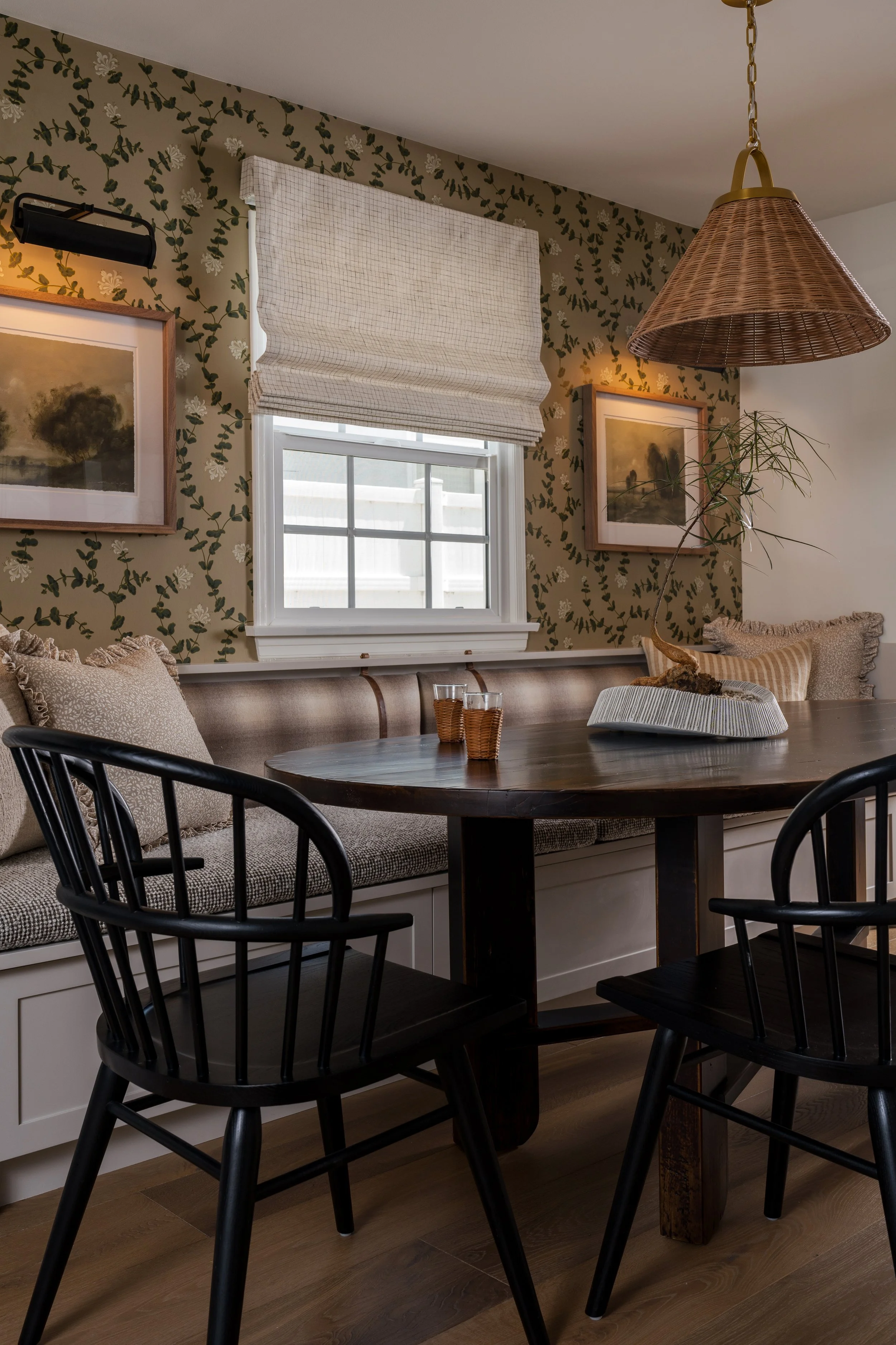 A cozy dining nook with a dark wooden round table, black chairs, a window with beige roman shade, floral wallpaper, and framed landscape artwork on the wall. There are beige cushions on the built-in bench and a hanging wicker pendant light.