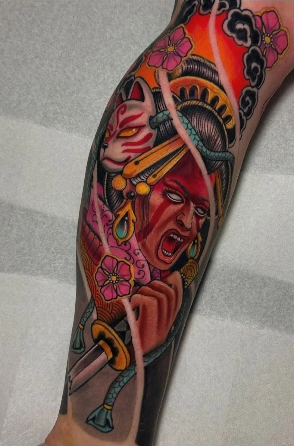 Tattoo of a traditional Japanese style featuring a woman with an angry expression, a white fox mask, cherry blossoms, a katana sword, and cloud motifs.