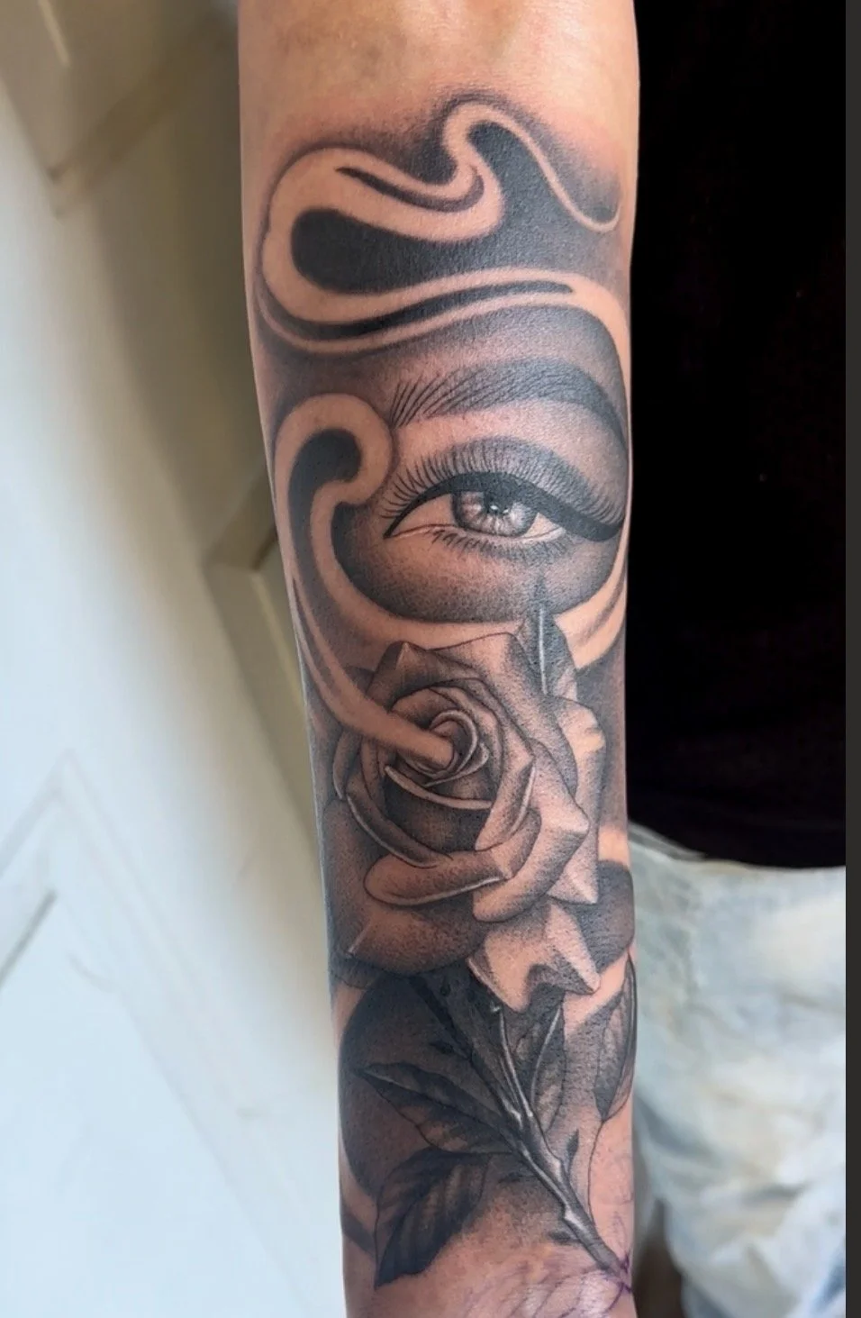 A detailed black and gray tattoo on a person's arm featuring a realistic eye, a rose, and swirling decorative elements.