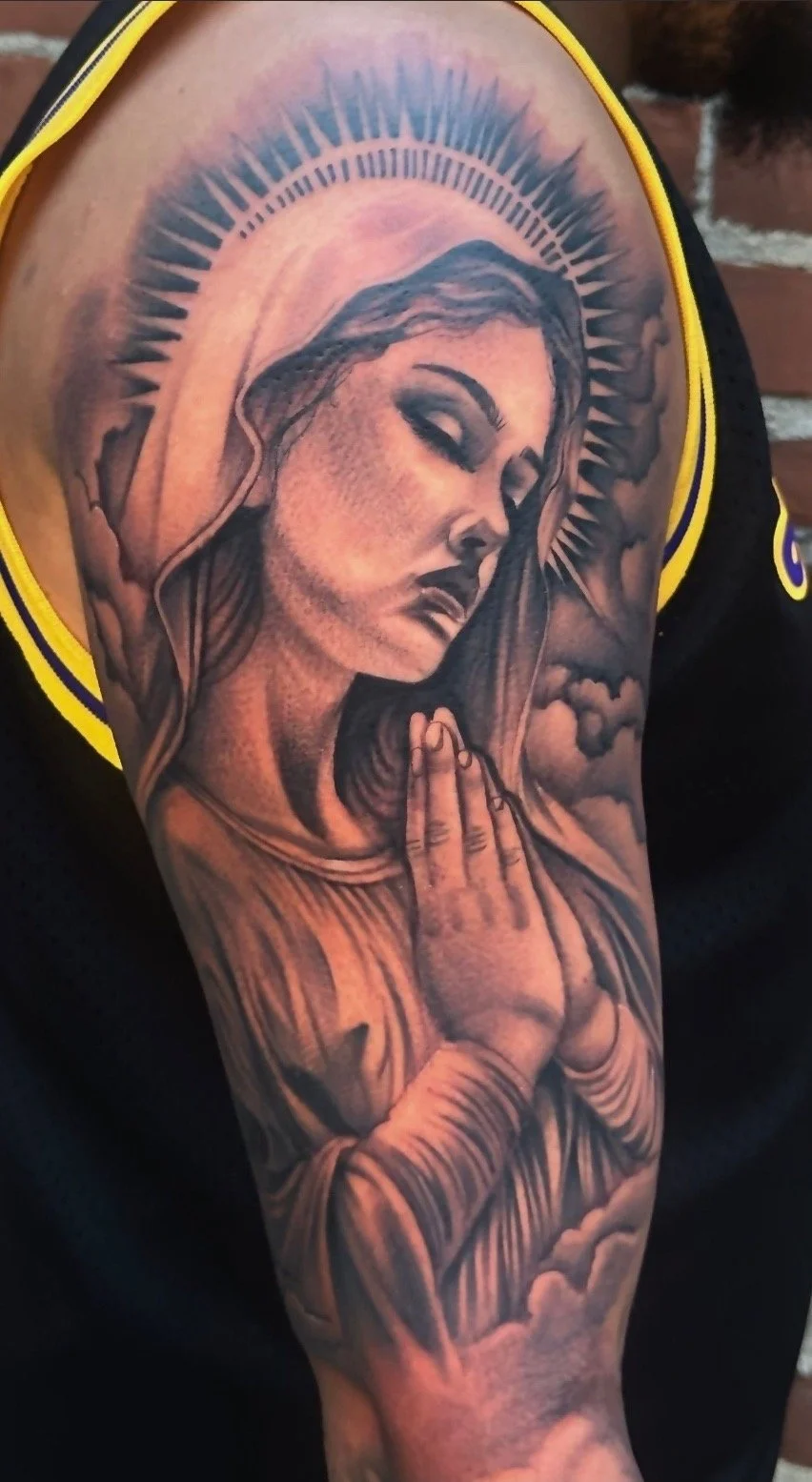 Tattoo of the Virgin Mary with hands in prayer, surrounded by a radiant halo and clouds.