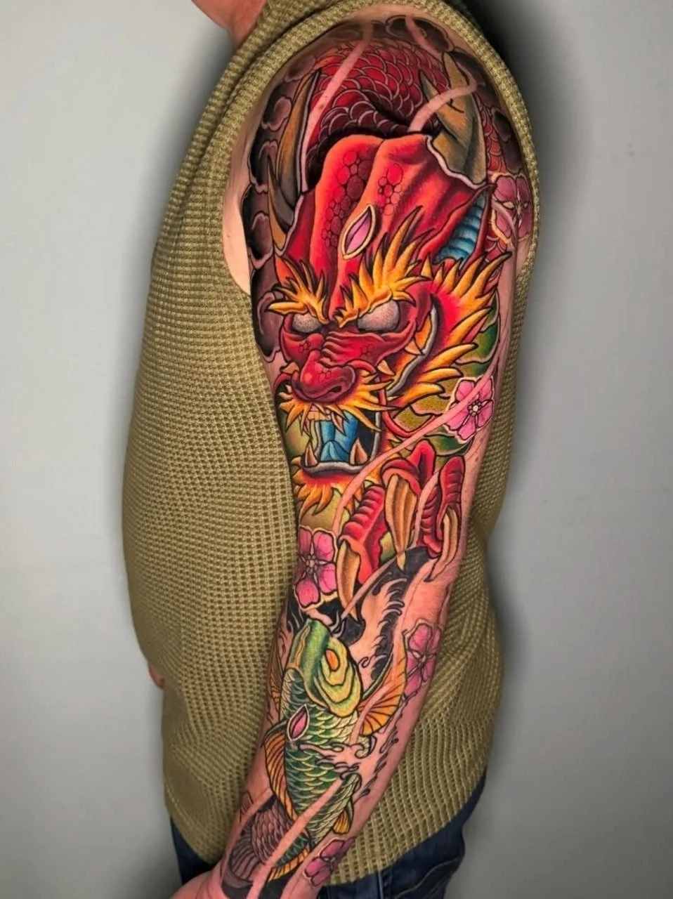 Colorful Japanese-style tattoo of a dragon on a person's arm, featuring vibrant reds, oranges, greens, and pinks with floral and wave details.