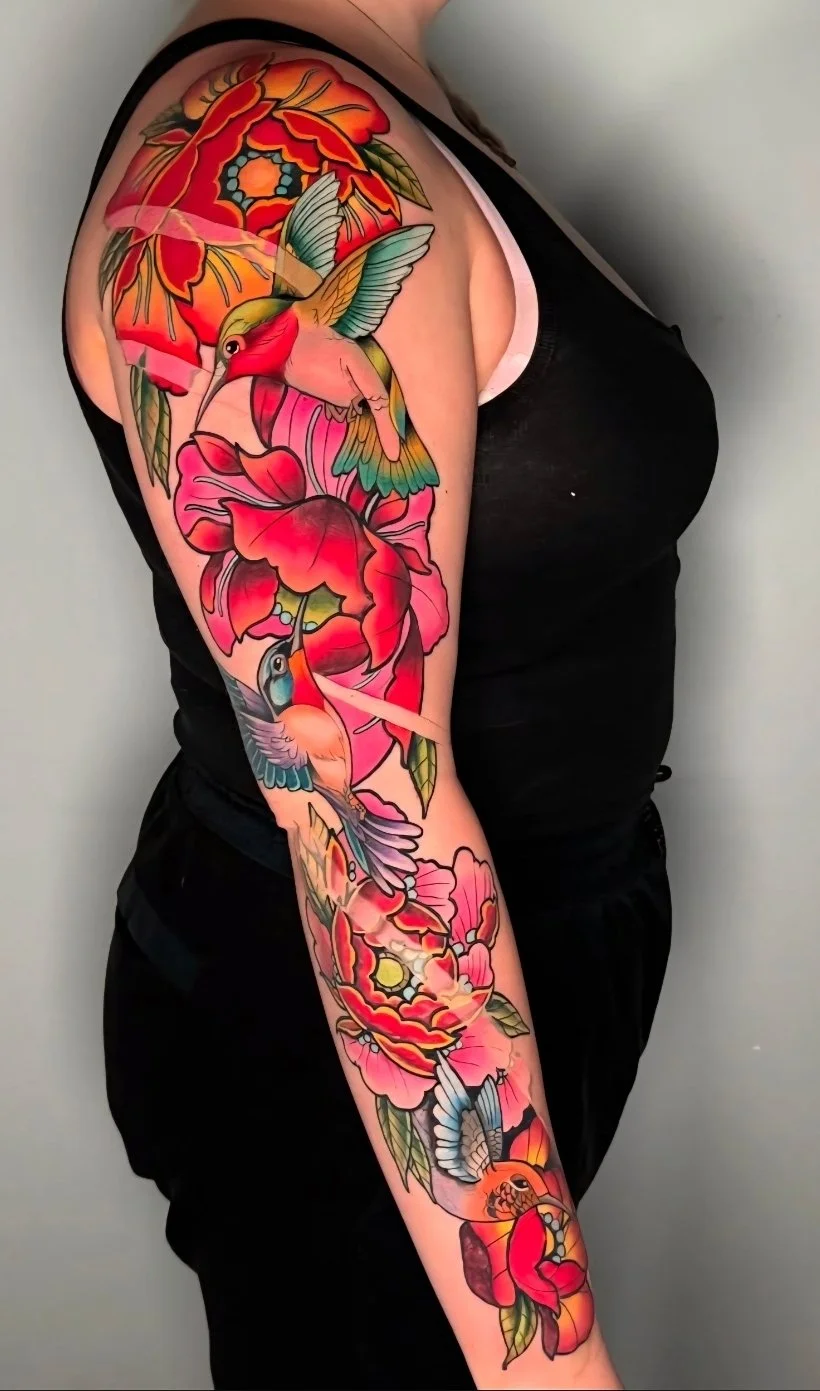 Colorful tattoo sleeve with hummingbirds, peonies, and abstract floral elements on a person's upper arm and forearm.