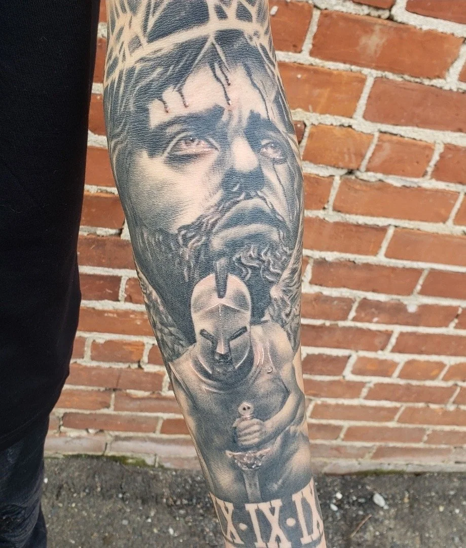 A black and gray tattoo on a person's arm depicting a serious male face with dreadlocks, holding a spray paint can.