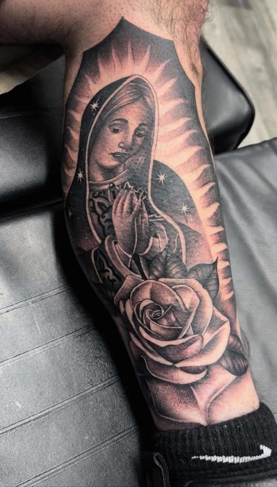 A detailed black and gray tattoo on a person's forearm depicting the Virgin Mary in prayer, with a halo of radiant light behind her, a large rose at the wrist, and stars around her head.