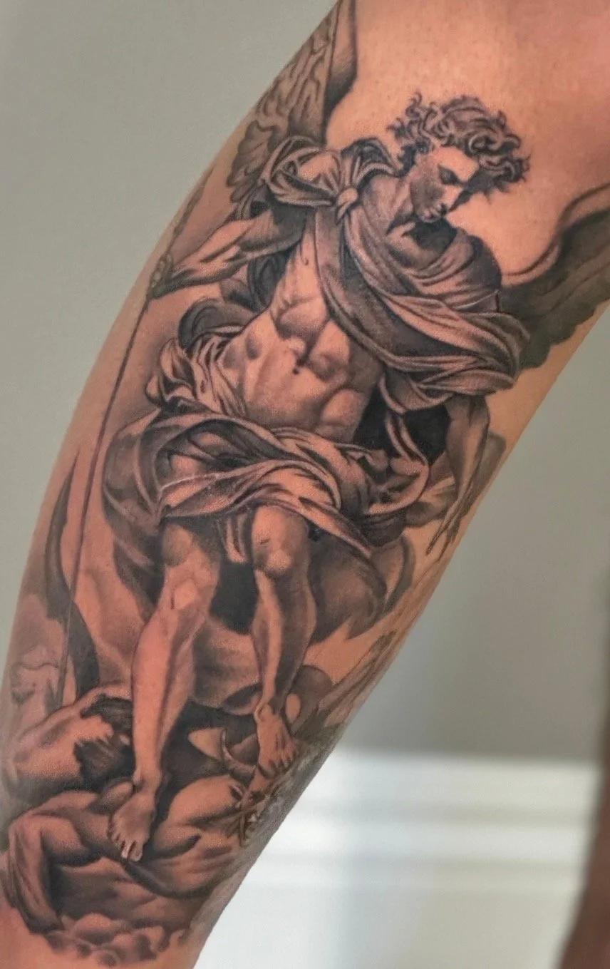 Black and gray tattoo of a muscular man with curly hair, wearing a flowing cloth, holding a spear, with clouds and a figure on a pile of rocks.