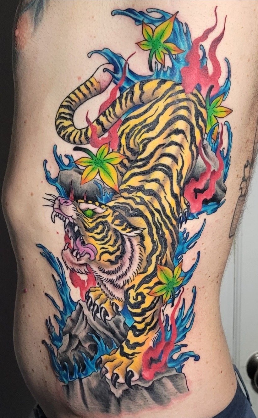 Colorful tattoo of a fierce tiger surrounded by blue and red flames and green leaves on a person's side.