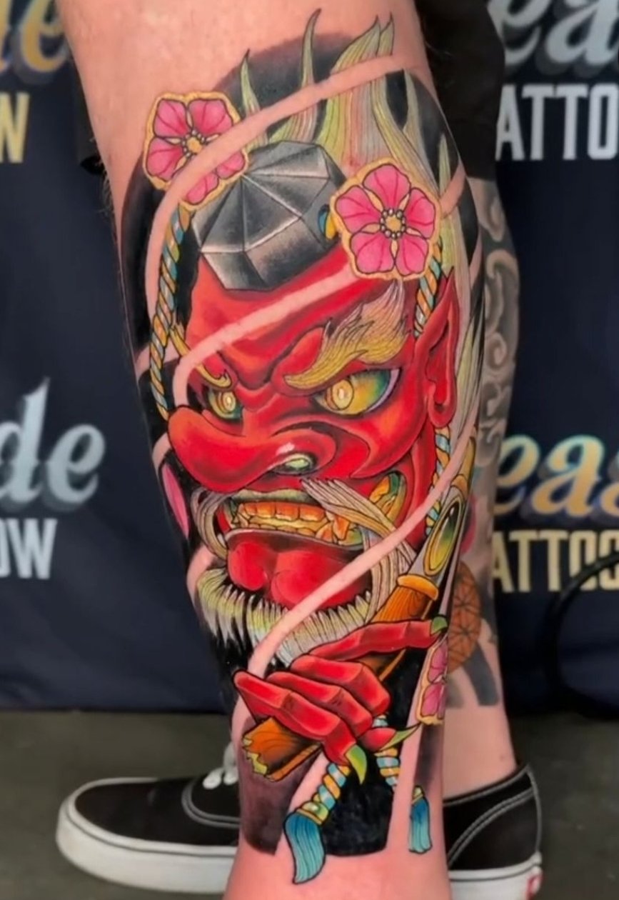 Colorful tattoo of a fierce red demon with yellow eyes, exaggerated facial features, and a long beard, holding a pipe, with cherry blossoms and decorative elements surrounding it.