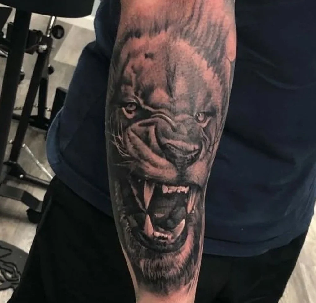 Tattoo of a fierce lion's face on a person's arm, showing the lion roaring with detailed shading.