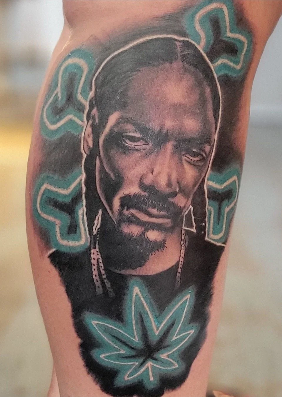 Black and grey portrait tattoo of Snoop Dogg with marijuana colored leaf and cross bones behind him.