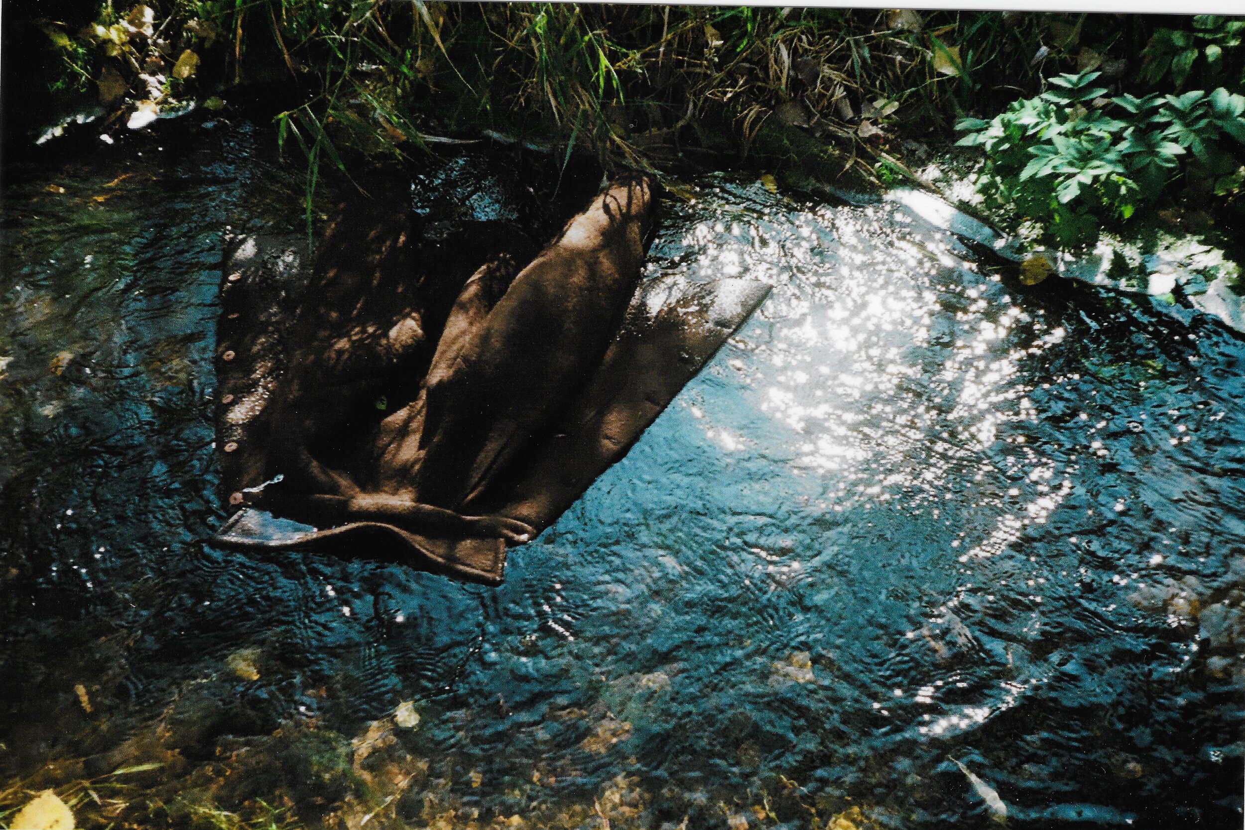 A brown glove submerged in a small, rocky stream with sunlight reflecting off the water surface.