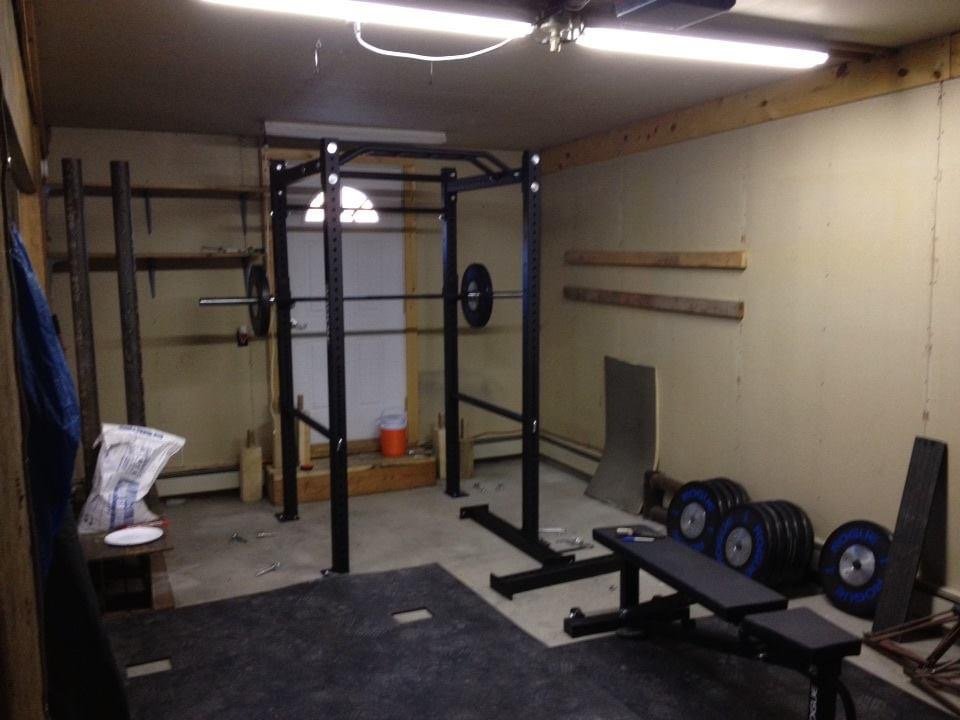 A home gym in a basement with a power rack, dumbbells, a weight bench, and workout equipment arranged along the walls.