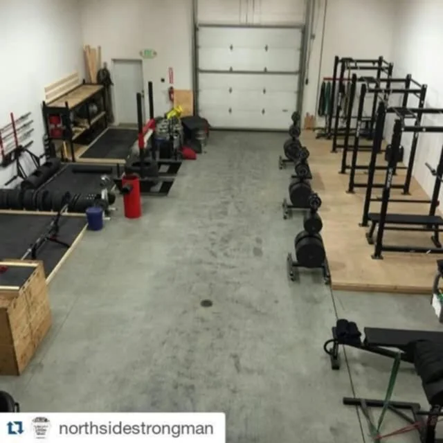 Empty gym or fitness training room with weightlifting equipment, barbells, squat racks, and benches, and a garage door at the back.
