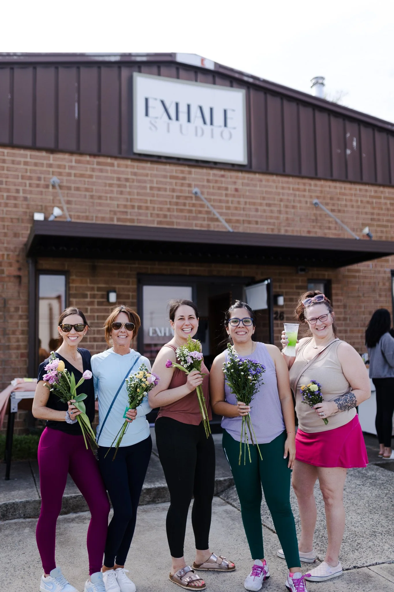 Group of five women holding flowers and drinks standing outside a brick building with a sign that reads 'Exhale Studio'