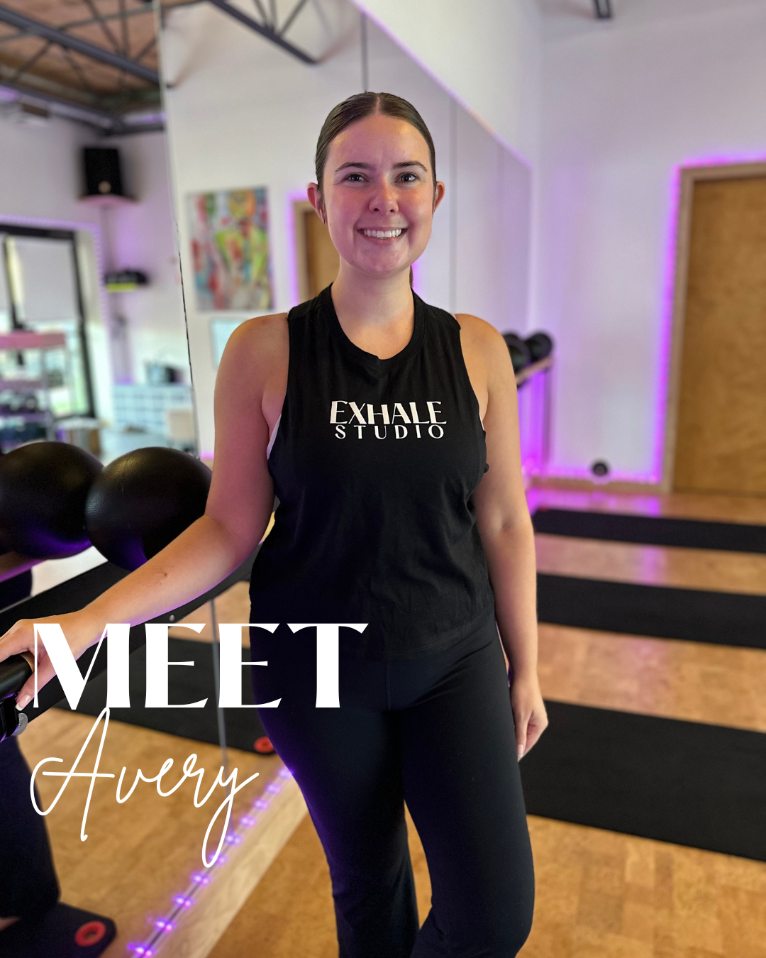 A woman standing in a fitness studio, smiling at the camera, wearing a black sleeveless top with 'EXHALE STUDIO' written on it, with black leggings. The studio has black mats and fitness equipment, with purple accent lighting.