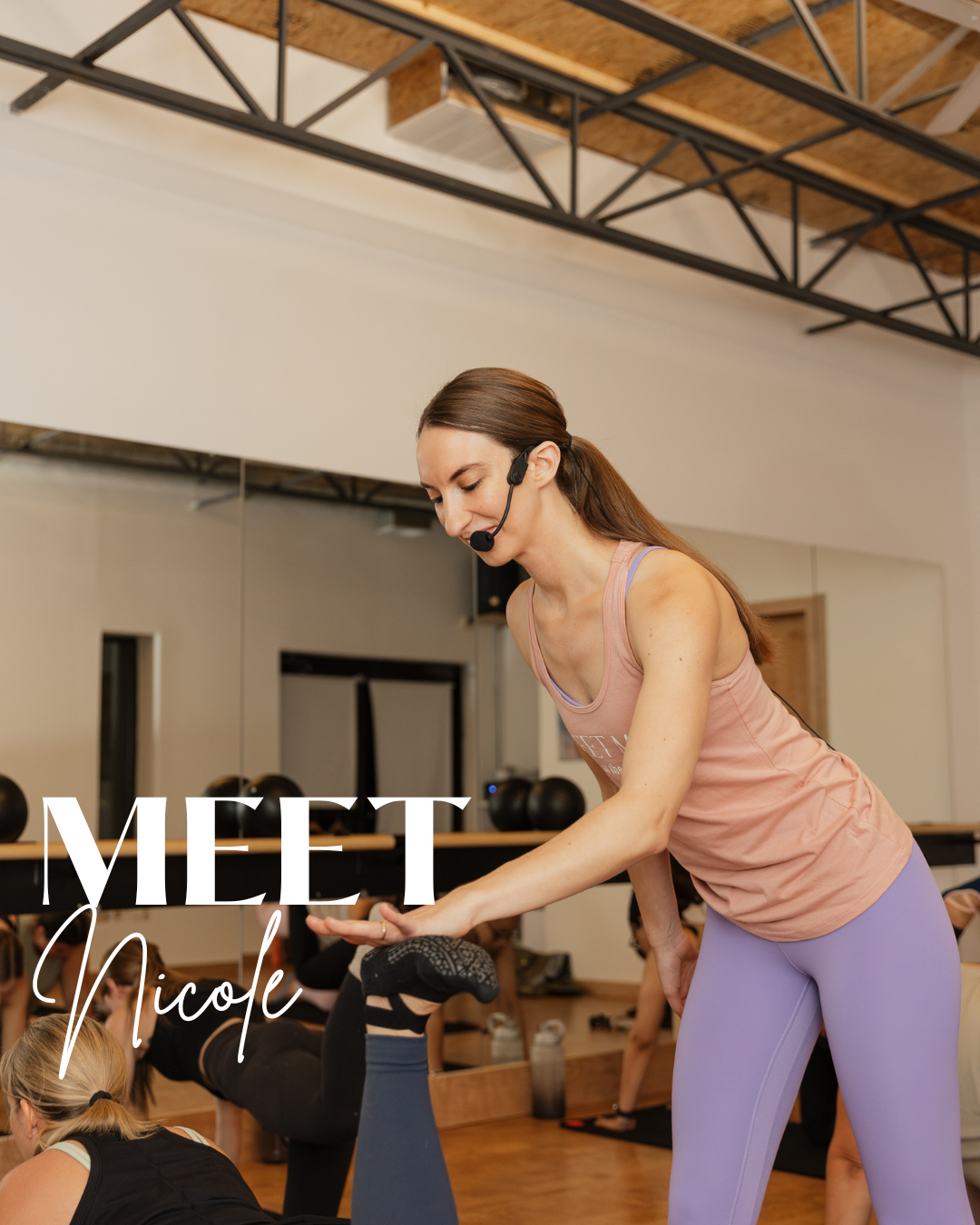 A fitness instructor with a headset assisting a person during a yoga class in a gym studio with wooden floors and wall-mounted mirrors.