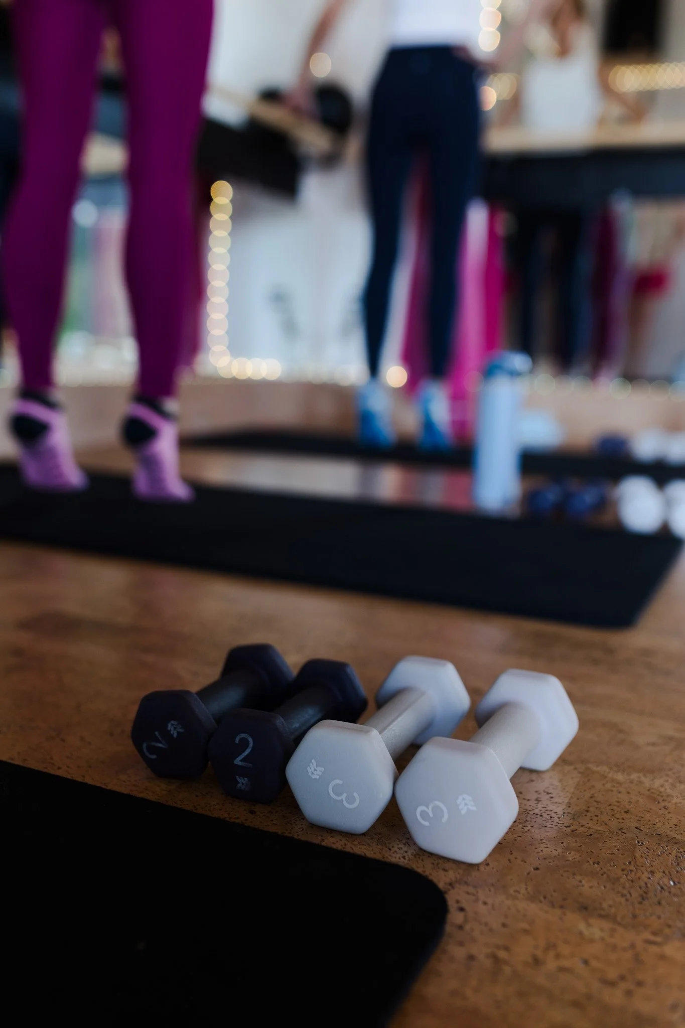A pair of black and white dumbbells with numbered weight labels, positioned on a wooden floor in a fitness class studio, with people exercising in the background.