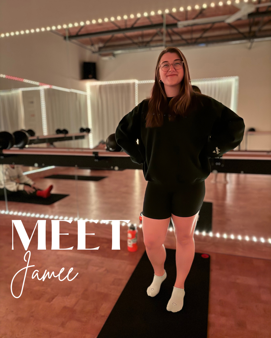 A young woman with glasses and long brown hair stands in a fitness studio, smiling confidently with her hands on her hips. She is wearing a black sweatshirt, black shorts, and white socks. The studio has a wooden floor, mirrors, and punching bags, and is decorated with string lights. The text overlays say "MEET Jame" in white and cursive font.
