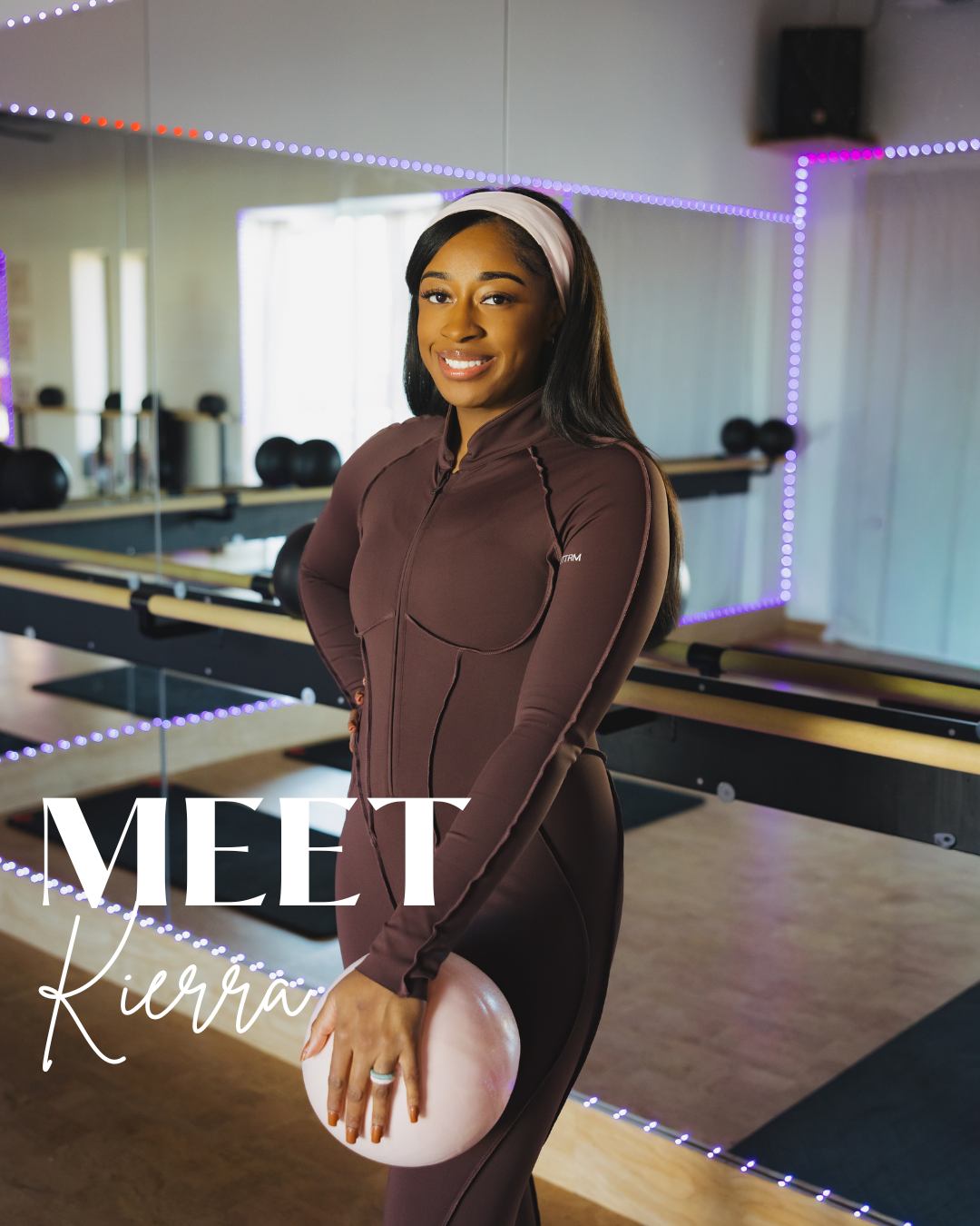 A woman in a brown athletic outfit standing in a fitness or dance studio, holding a pink exercise ball, with a large mirror and purple LED lights in the background.