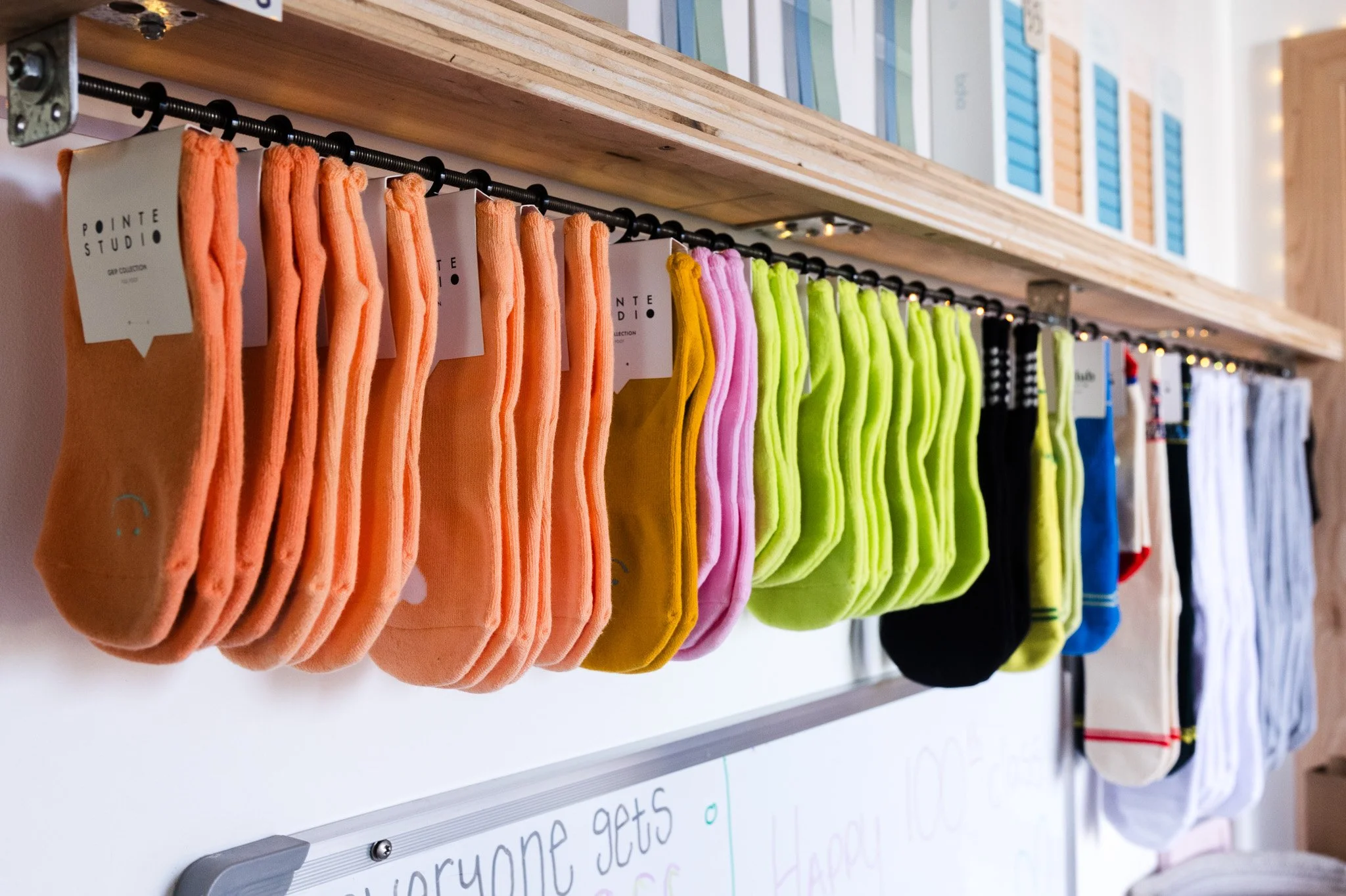 Colorful socks hanging on a rack inside a store, arranged by color from peach to white, with a shelf above and a whiteboard below.